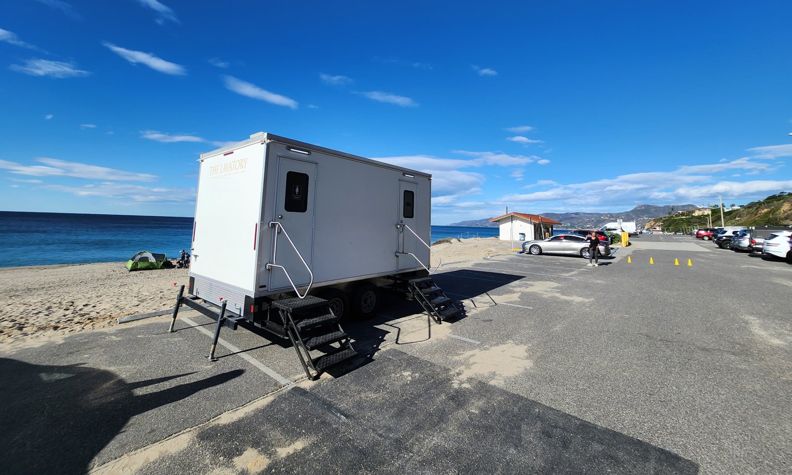 2-station shower and restroom combo trailer with its stairs set on a beach parking lot next to the sand, the ocean and coastal hills behind it