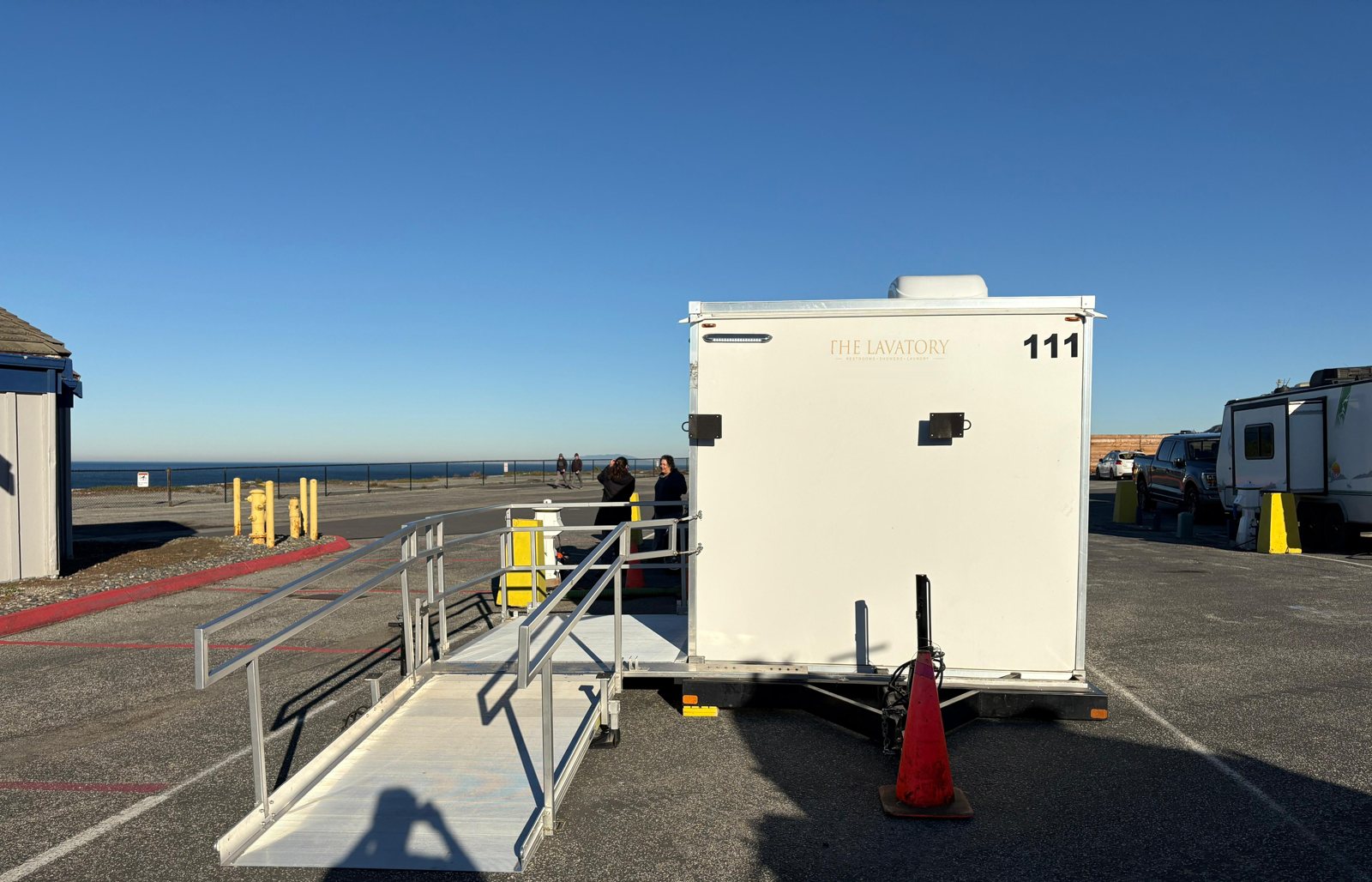 ADA shower trailer seen from the rear with an aluminum ramp and rails set on a coastal parking lot, the ocean and a fenced bluff walkway behind it