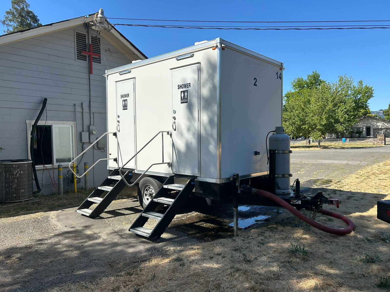 2-station shower and restroom combo trailer with two stair units, a propane tank on the tongue and a red sewer hose connected, parked beside a gray building on dry grass