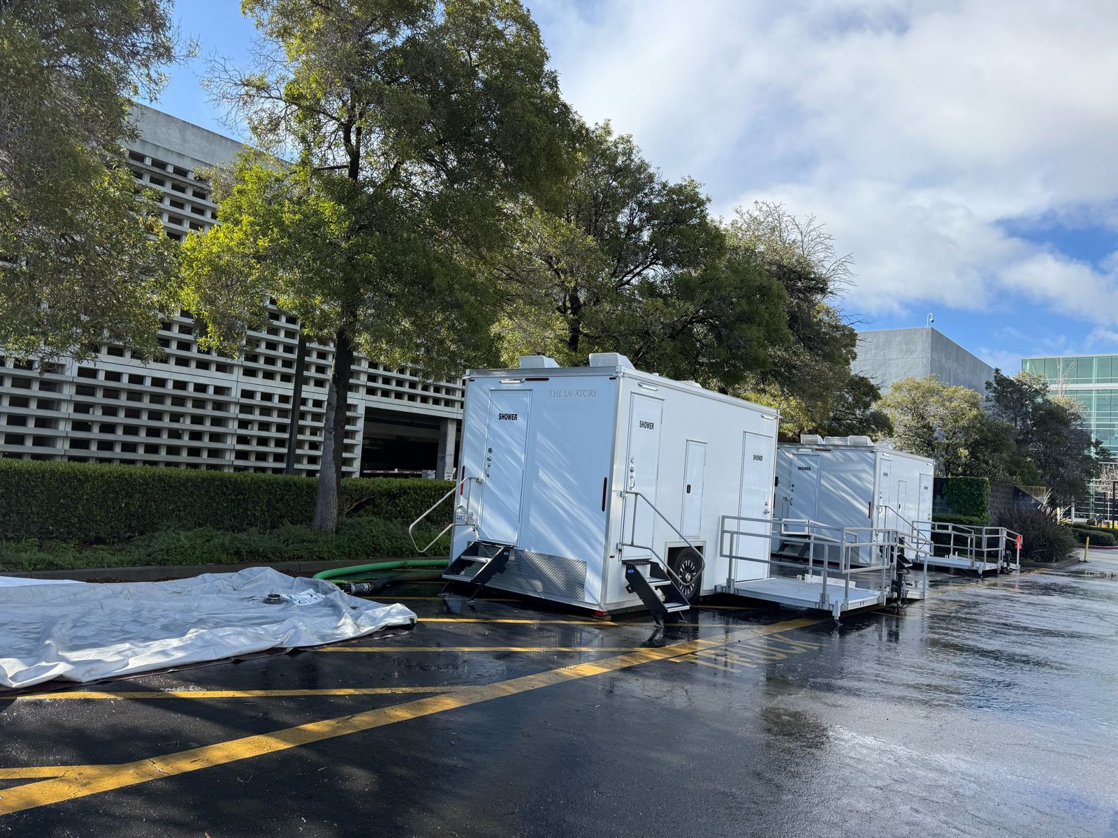 8-station private shower trailer and an ADA shower trailer with its ramp set on a wet business park parking lot, oak trees and a concrete parking structure behind them