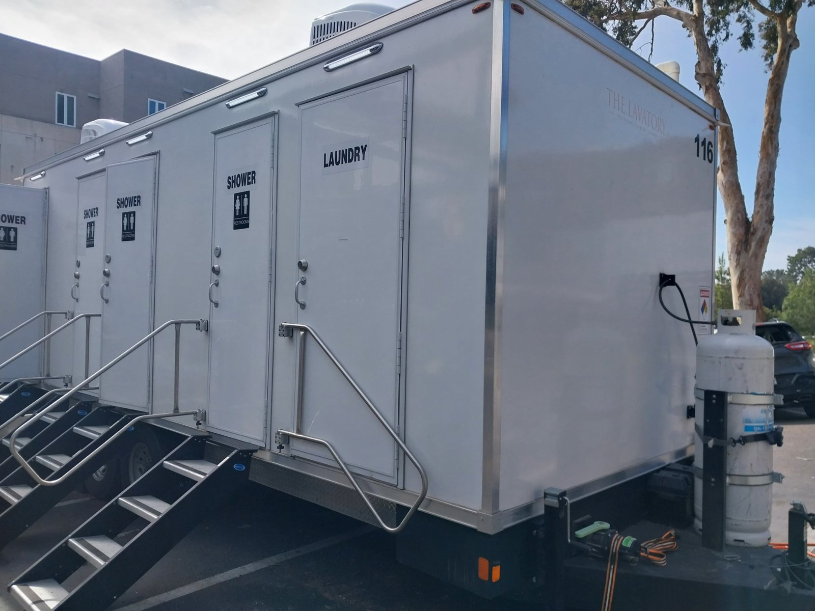 4-station shower, restroom and laundry combo trailer with SHOWER and LAUNDRY doors and stairs, parked in a lot beside a building and a eucalyptus tree