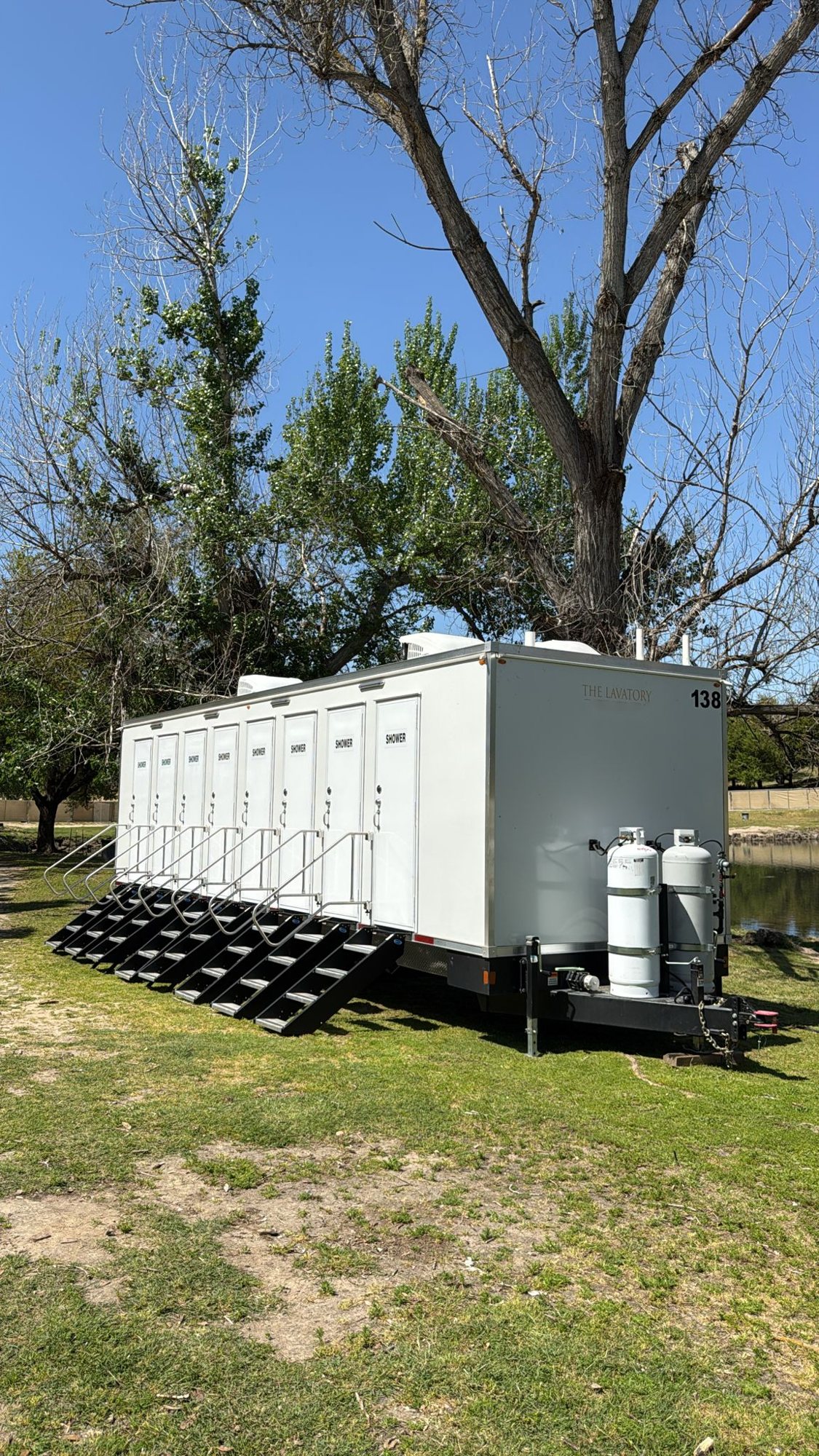 8-station private shower trailer with a stair at each of its eight SHOWER doors, parked on a park lawn under cottonwood trees beside a pond