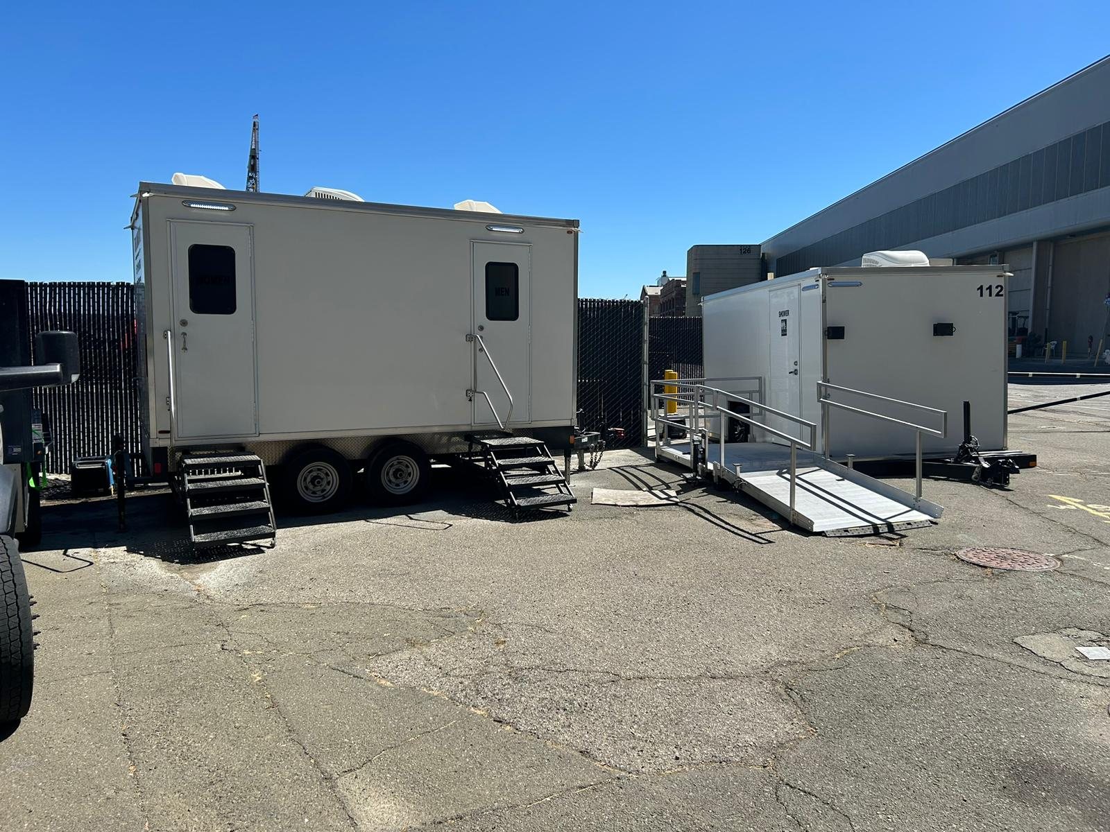 Two-room restroom trailer next to an ADA shower trailer with its aluminum ramp, both parked in a fenced industrial yard beside a large warehouse building