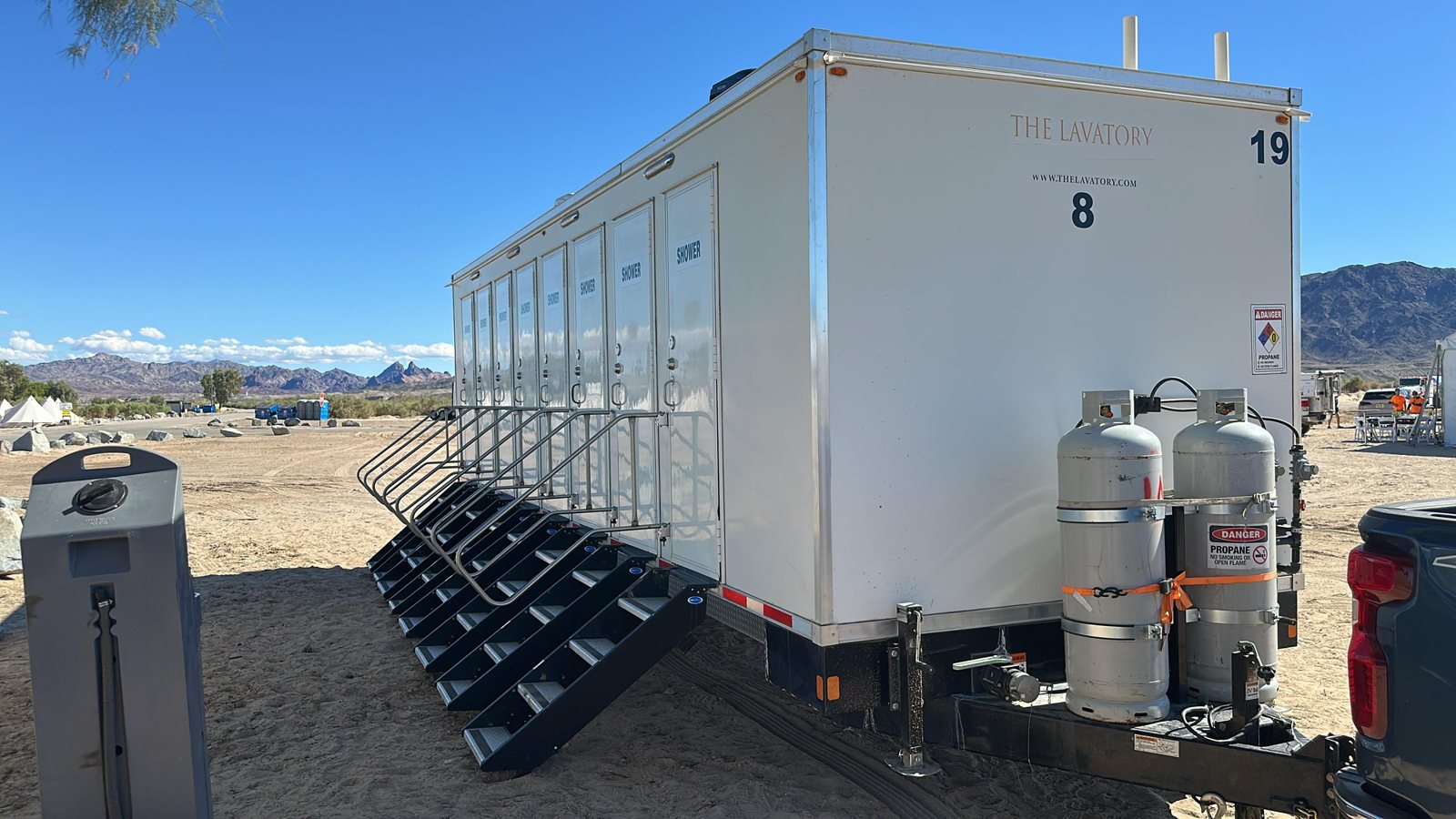 8-station private shower trailer with eight doors and eight stair units set up on a sandy lot with propane tanks on the tongue and dry hills behind it