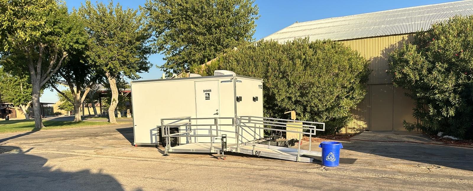ADA shower trailer with a railed ramp and landing on a paved lot beside shade trees and a metal building