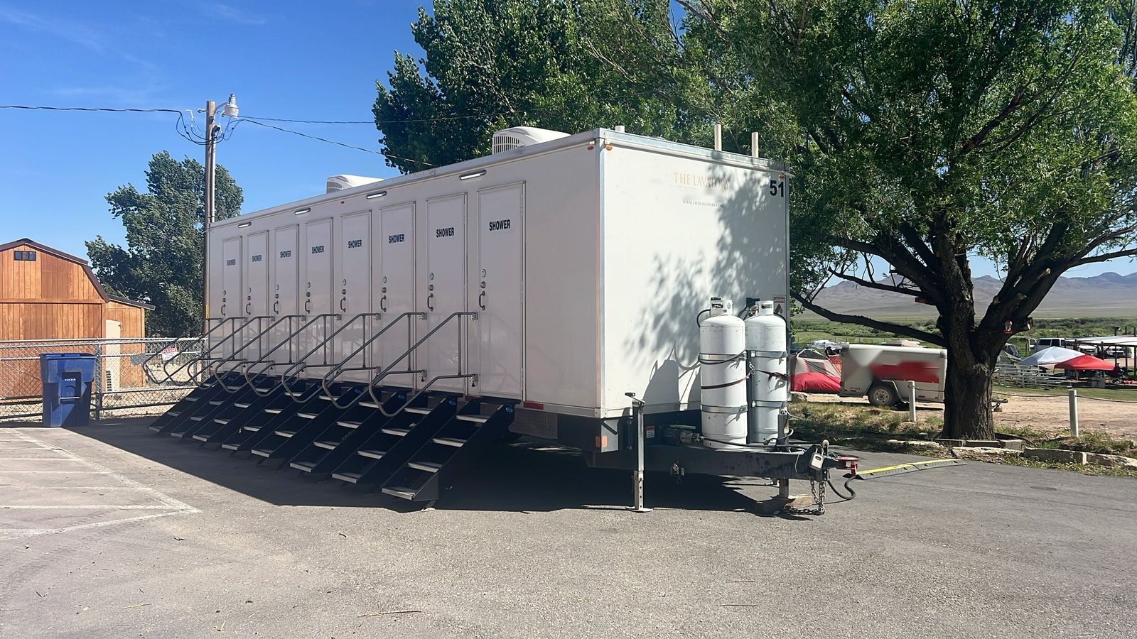 8-station private shower trailer with eight stair units on a paved lot beside a campground with tents, shade trees and a wooden barn