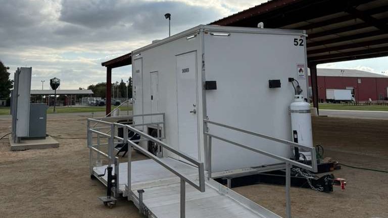 ADA + 2 shower trailer with its aluminum ramp and rails set up on dirt beside a covered fairground pavilion