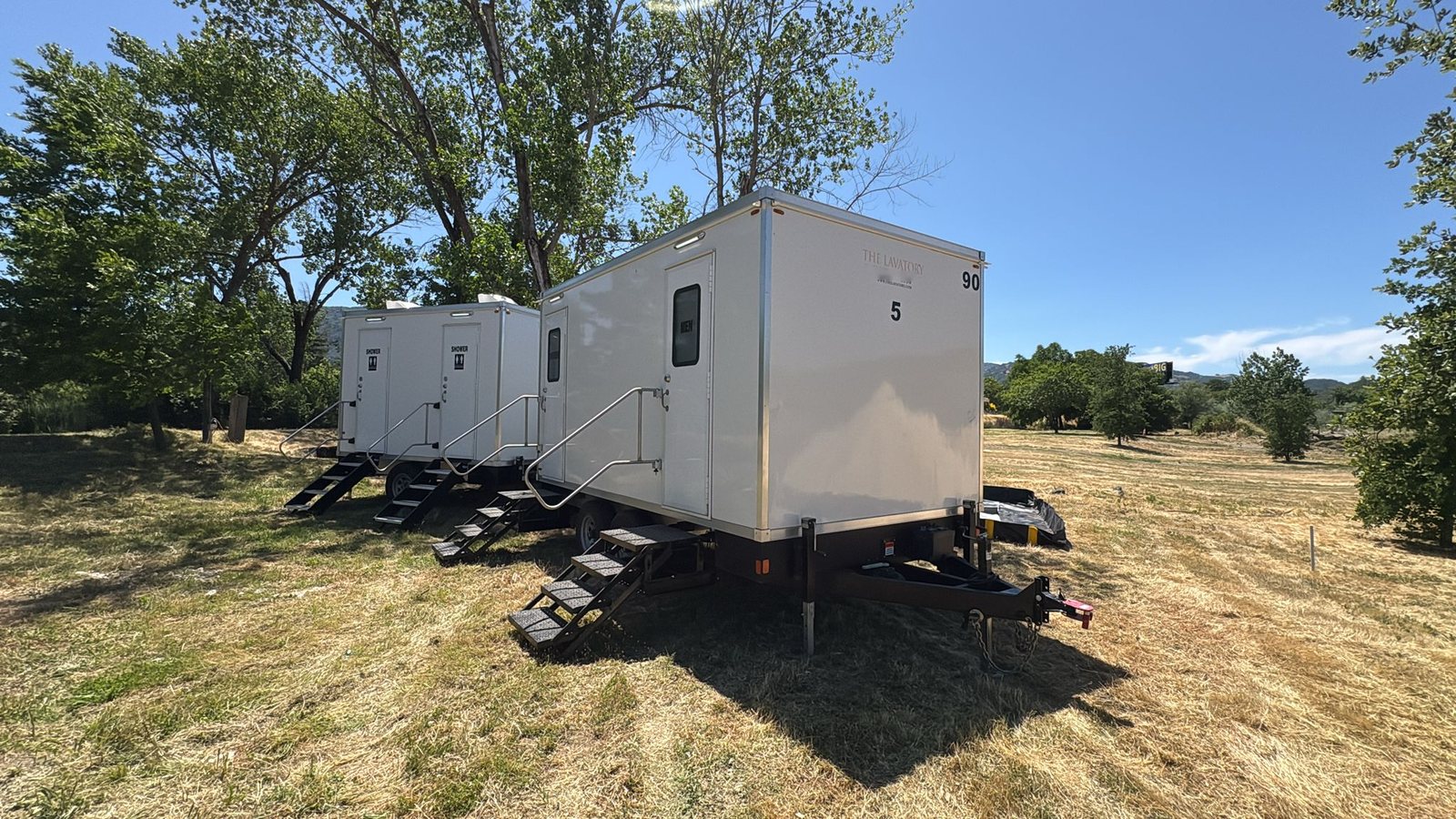 2-station shower and restroom combo parked behind a restroom trailer on a dry grass field under tall shade trees