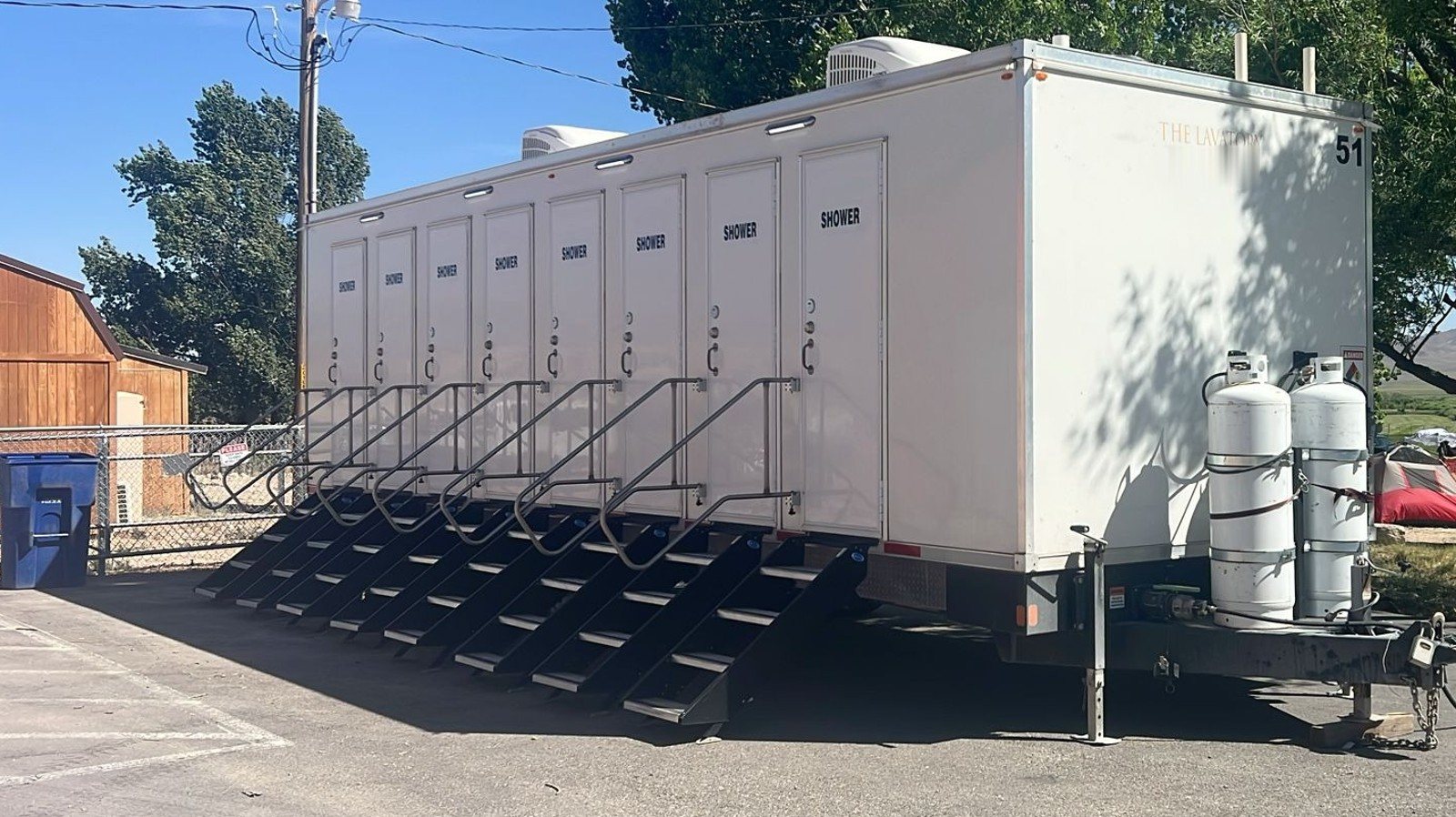 8-station shower trailer with eight doors and eight black stair units parked on a paved lot under shade trees, twin propane tanks on the tongue