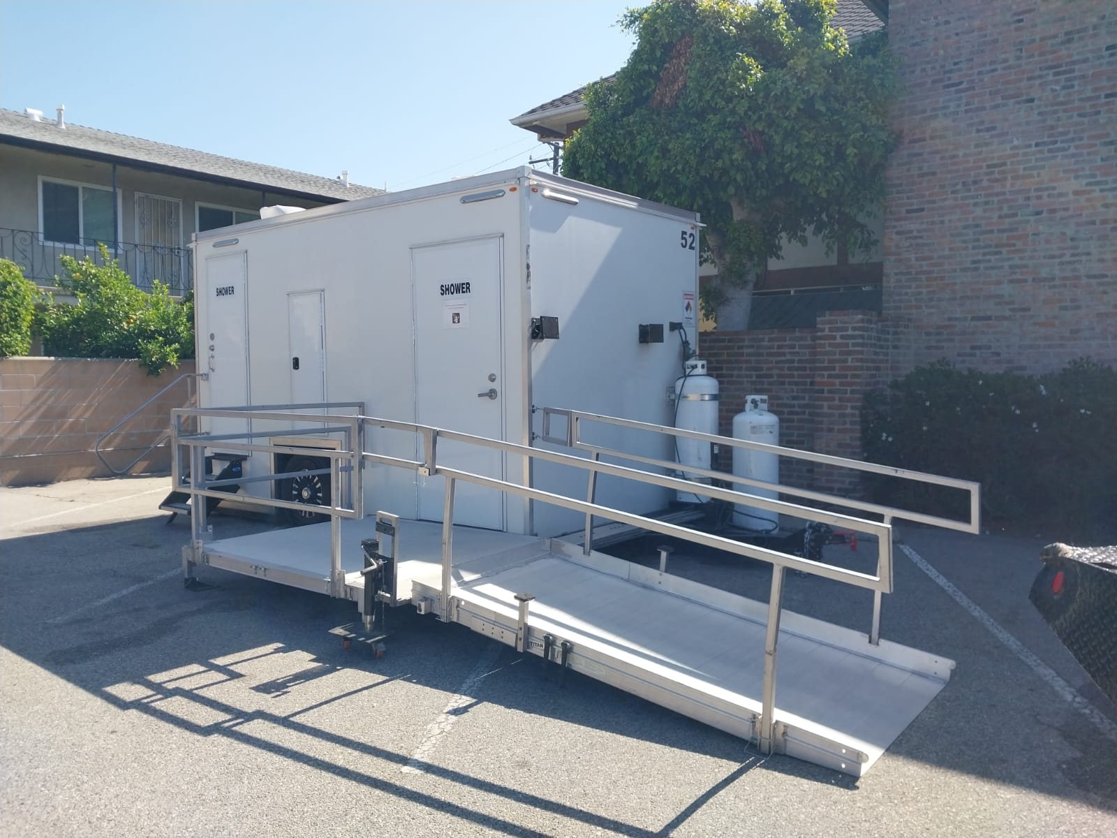 ADA plus two shower trailer with a railed aluminum ramp in a paved lot between an apartment building and a brick wall