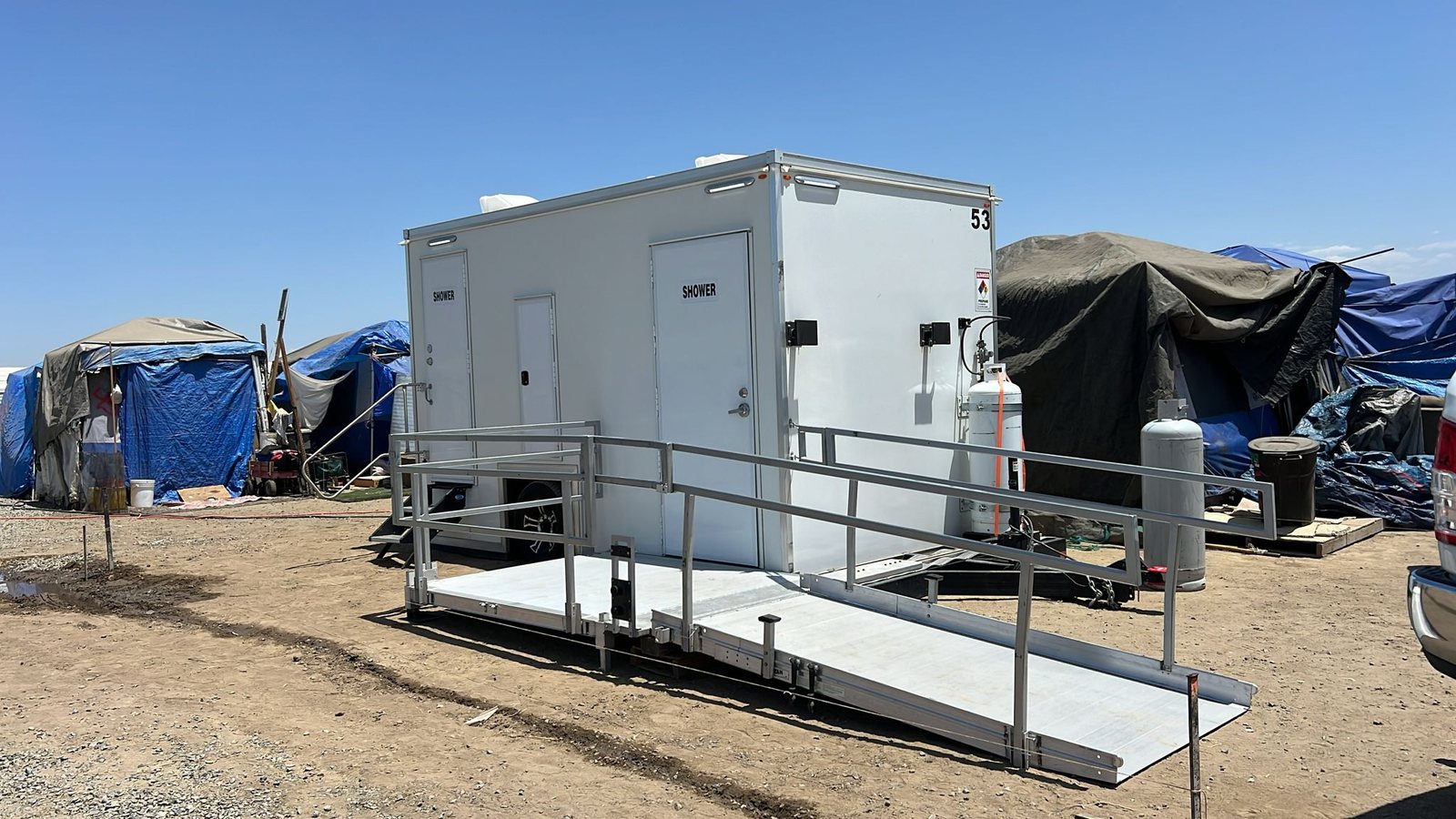 ADA plus two shower trailer with its own ramp and propane tanks set on bare dirt beside tarp-covered shelters, no hookups nearby