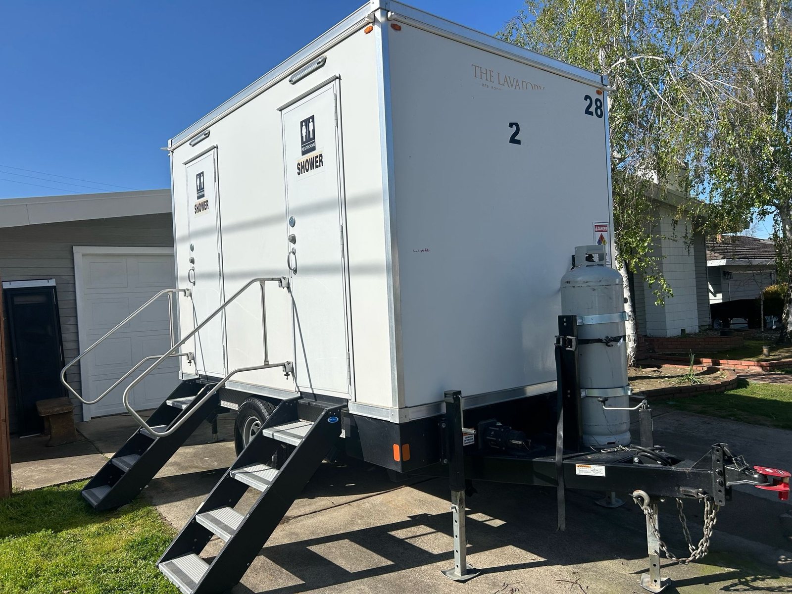 2-station shower and restroom trailer with two stair units parked on a home driveway beside a garage