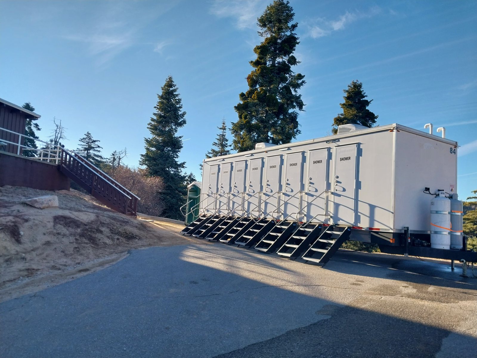 8-station shower trailer with eight doors and eight stair units on a paved lot among tall pines, propane tanks on the tongue