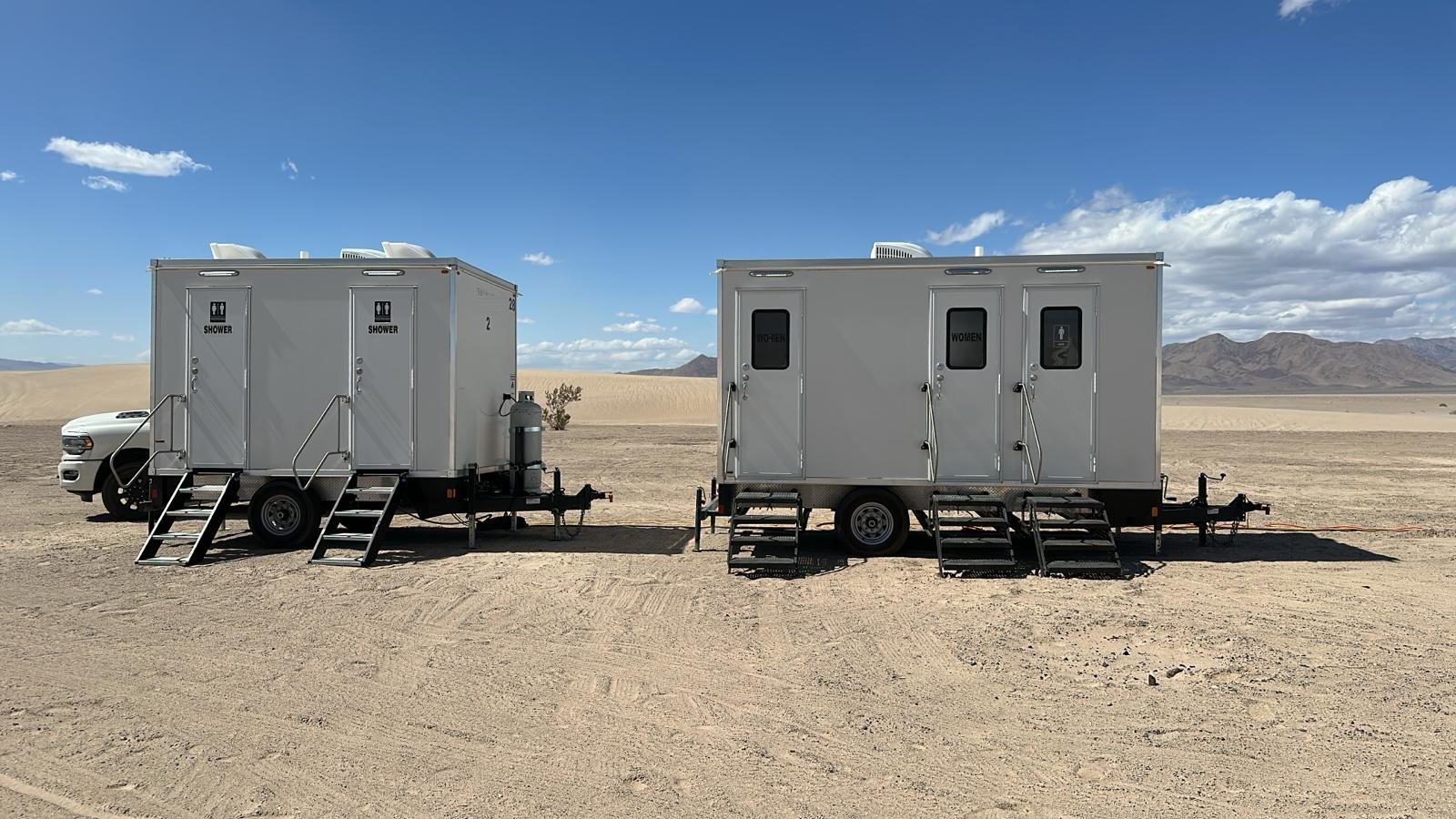 Two shower trailers parked on open desert ground with mountains behind and a pickup truck beside the first unit