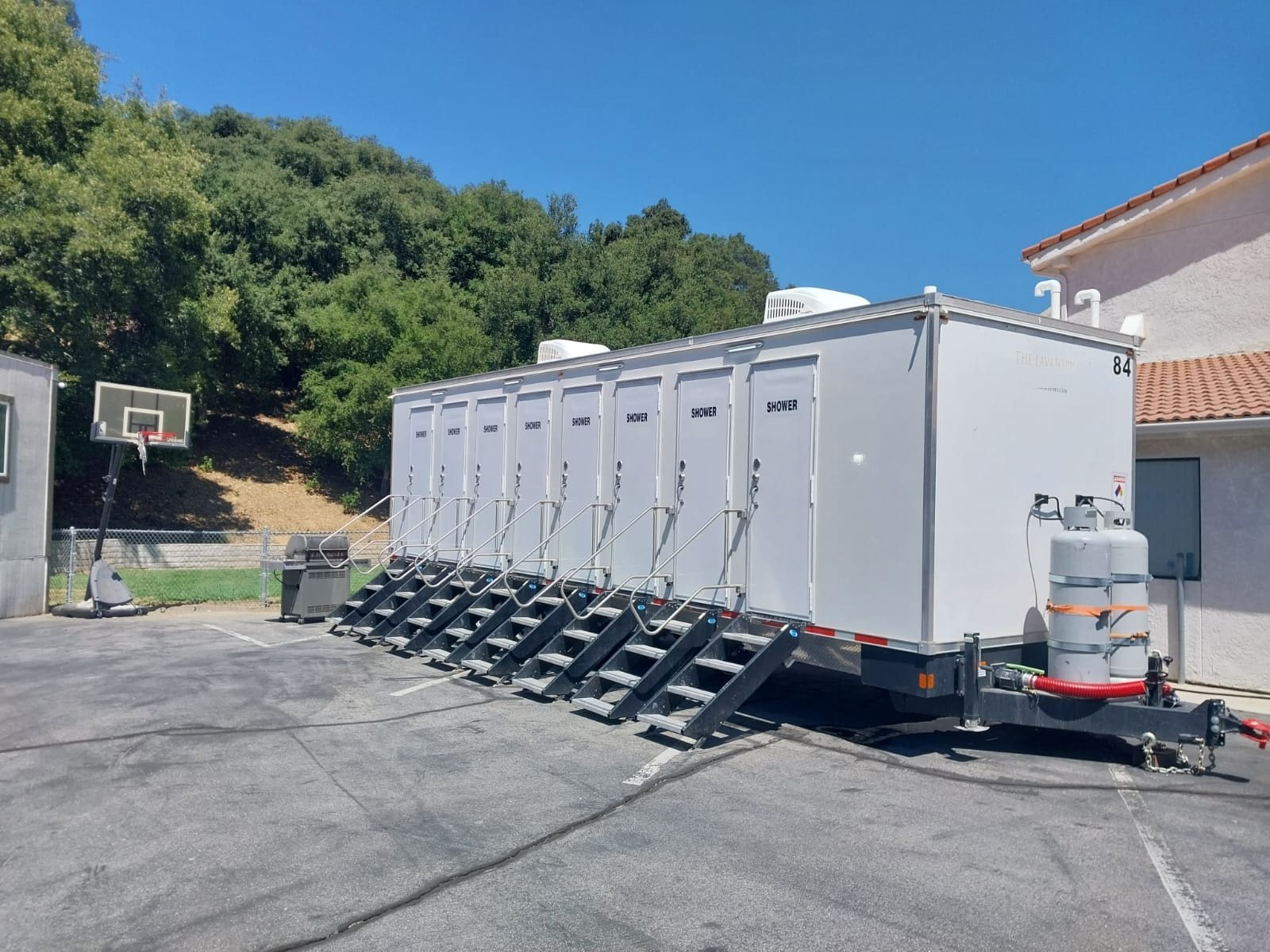 8-station shower trailer with eight shower doors and stair units on a paved lot next to a basketball hoop and an oak-covered hillside