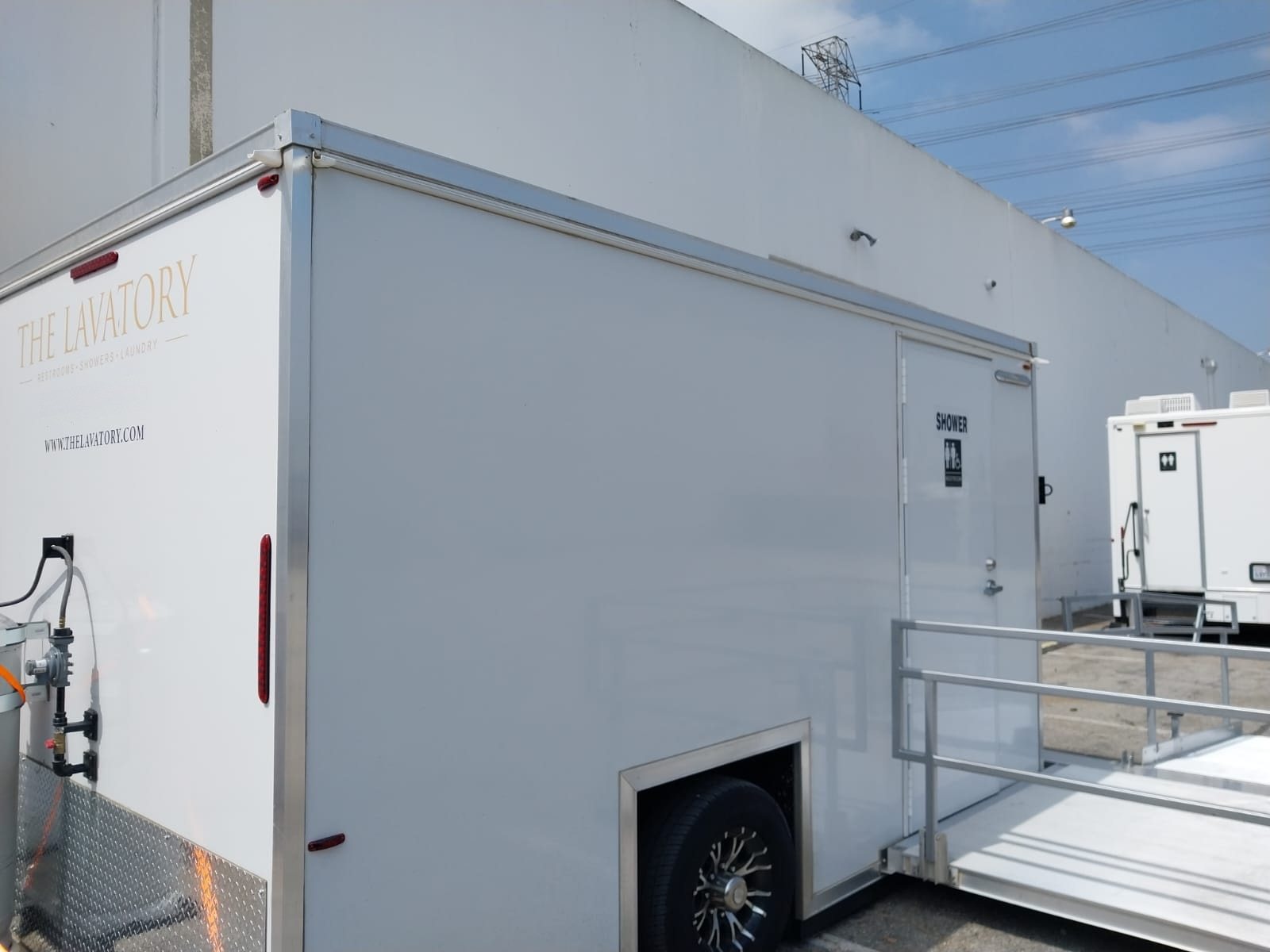 ADA plus two shower trailer with its railed ramp set up on asphalt beside a tall concrete warehouse wall, power lines overhead