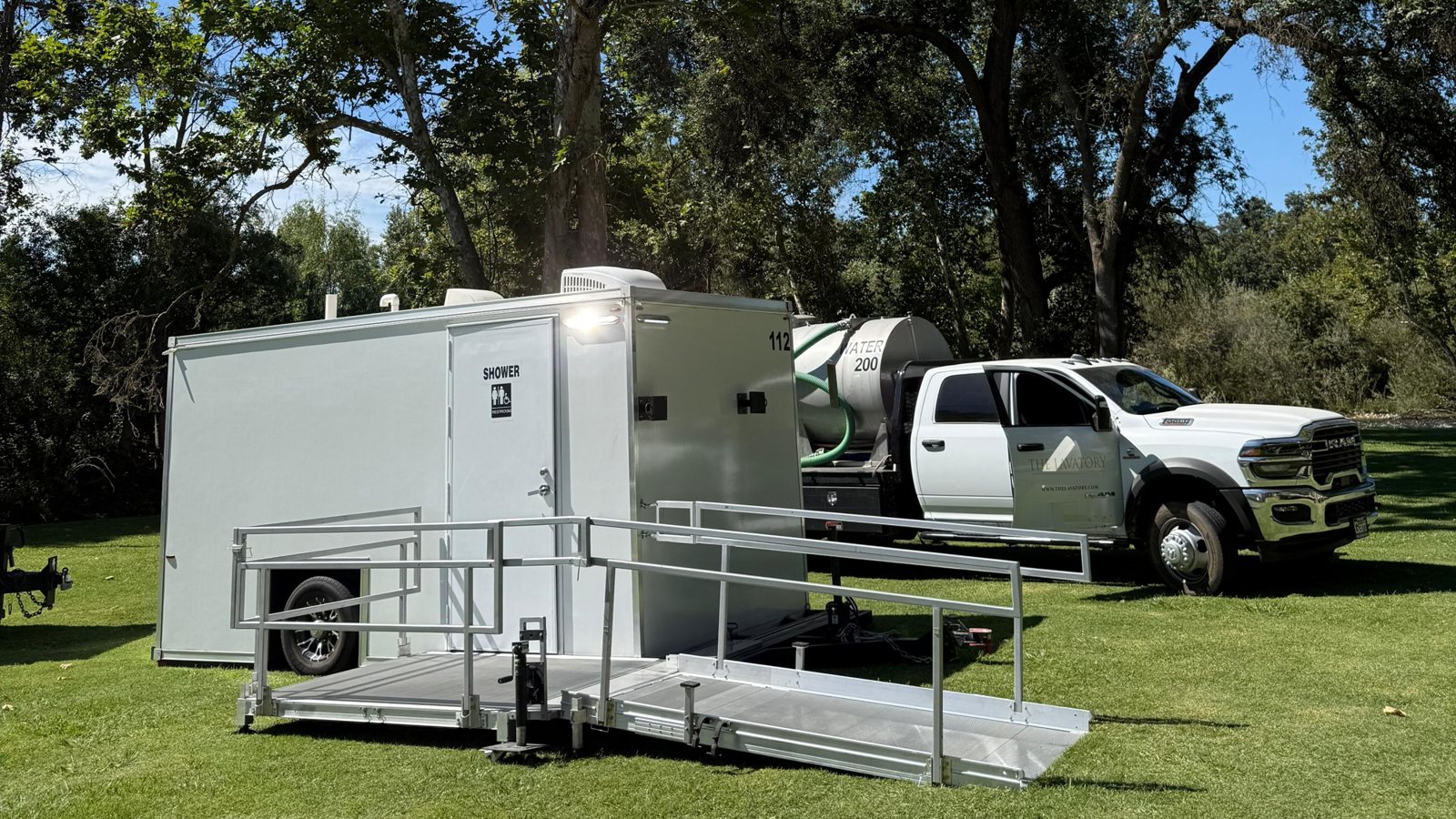 Single-station ADA shower and restroom combo trailer with its railed ramp set up on a park lawn, a service truck carrying a water tank parked behind it
