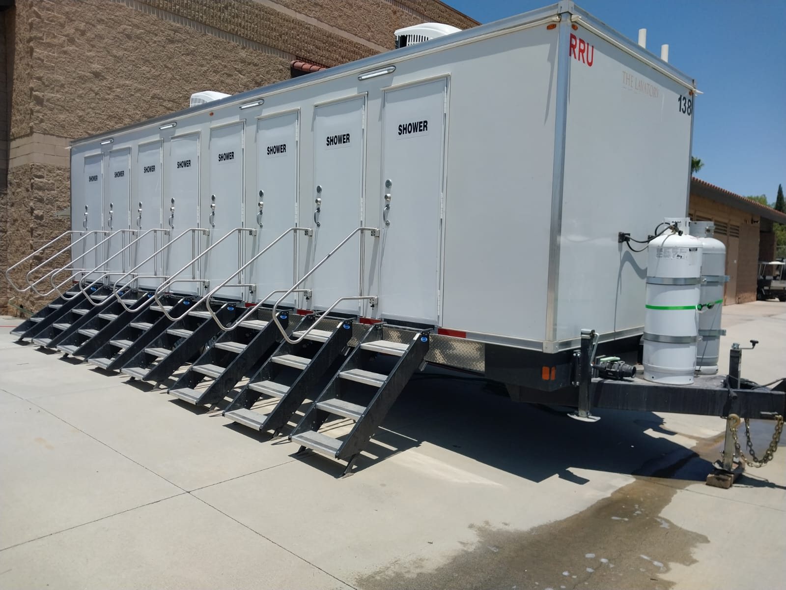 8-station shower trailer with eight stair units set on a concrete lot beside a block building, propane tanks on the tongue