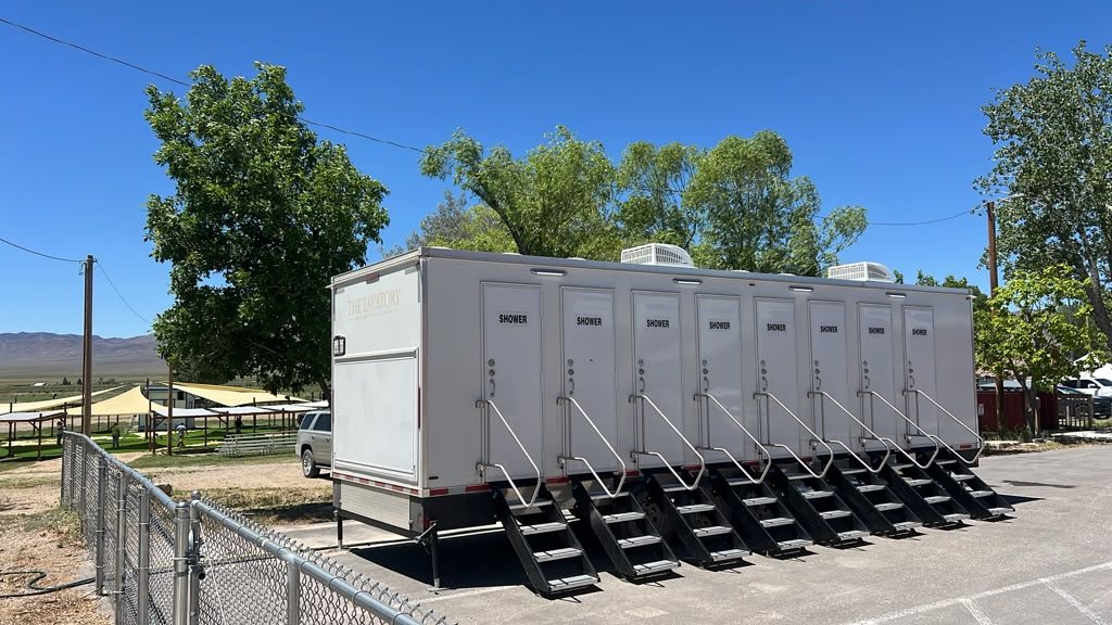 8-station shower trailer with stairs at every door on a paved lot behind a chain link fence, shade canopies and hills in the distance
