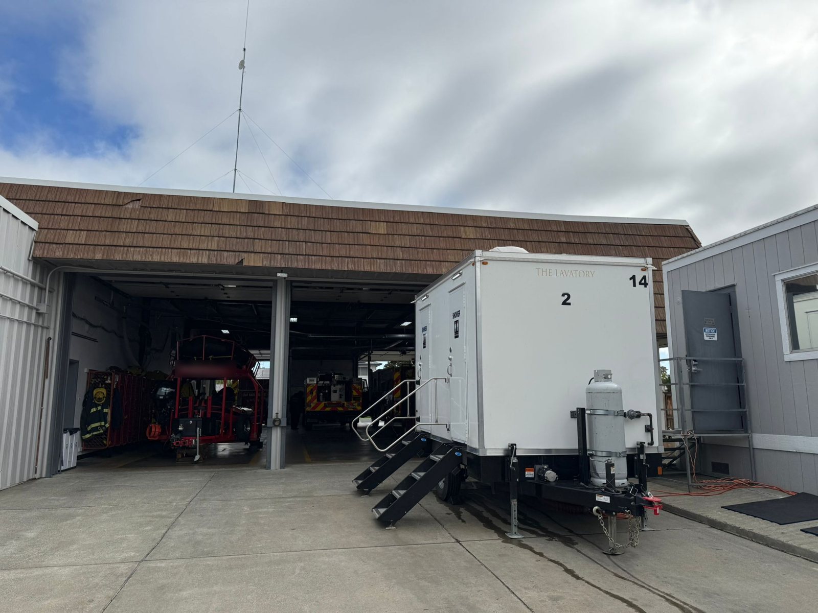 Two-room shower and restroom trailer parked on the apron beside an open fire station bay, with turnout gear and a fire engine inside