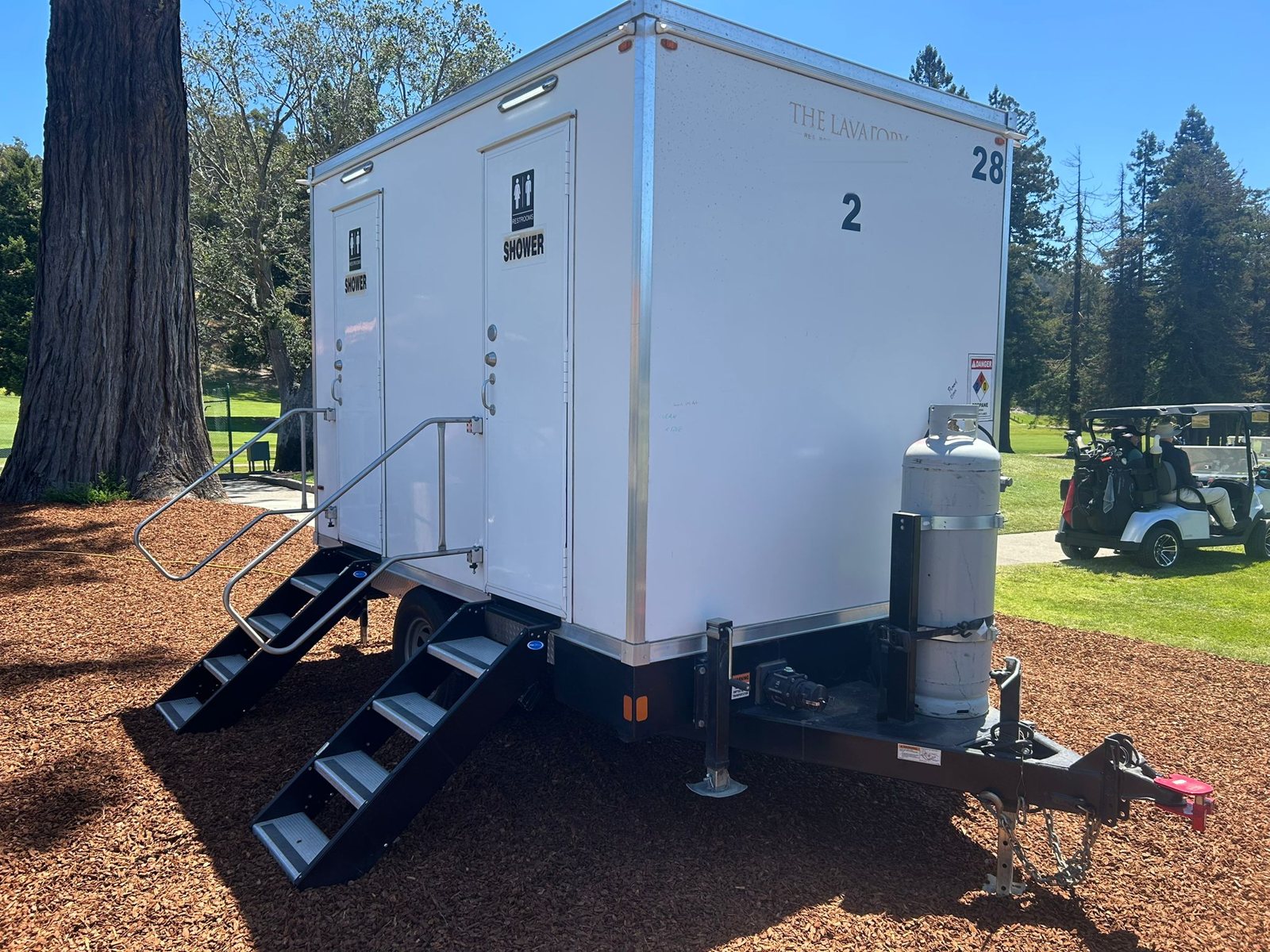 Two-room shower and restroom trailer with folding stairs set on bark mulch beside a golf course, a golf cart passing on the cart path