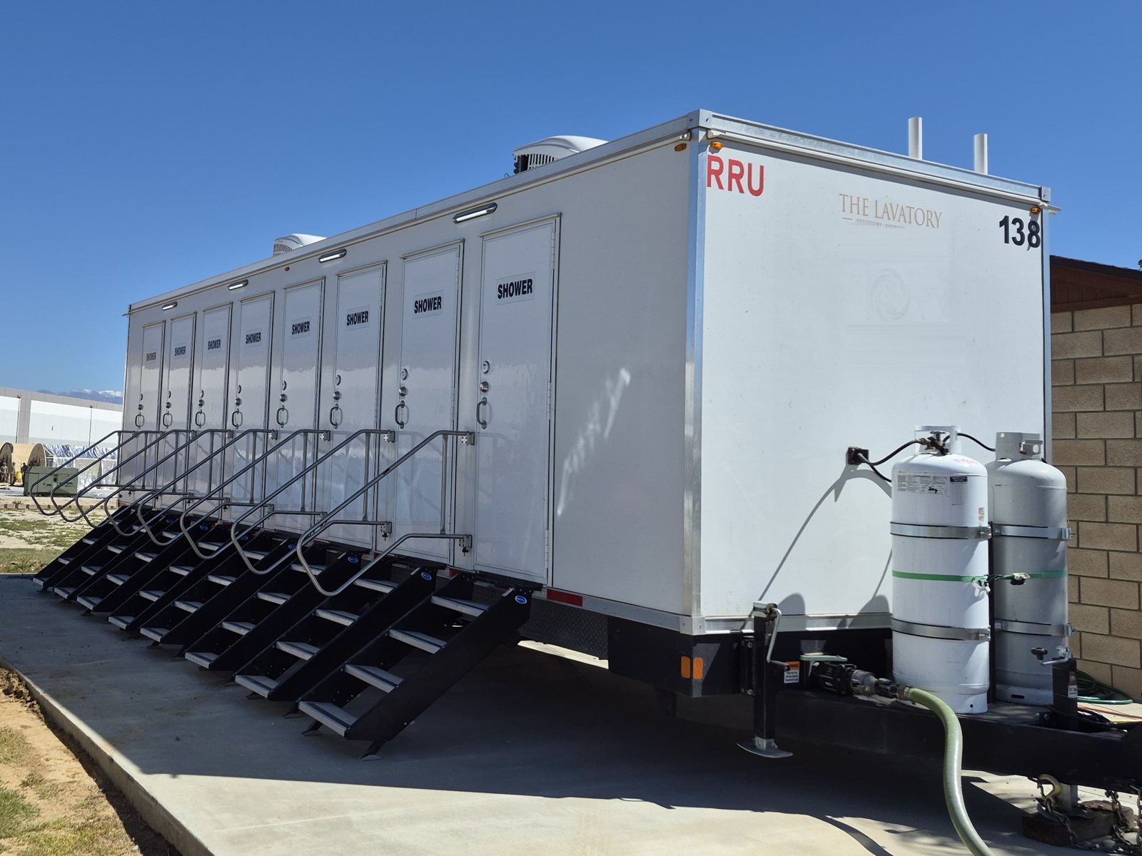 8-station private shower trailer with eight doors and eight stair units on a concrete pad, propane tanks at the tongue and large warehouse buildings in the distance