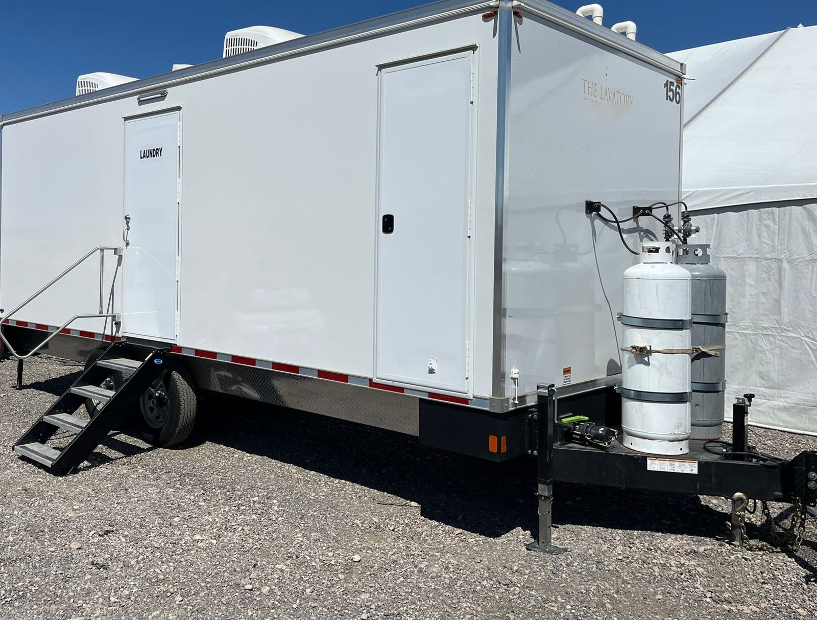 4-station shower, restroom and laundry combo trailer with a LAUNDRY door and stairs, parked on a gravel work yard beside a white tent