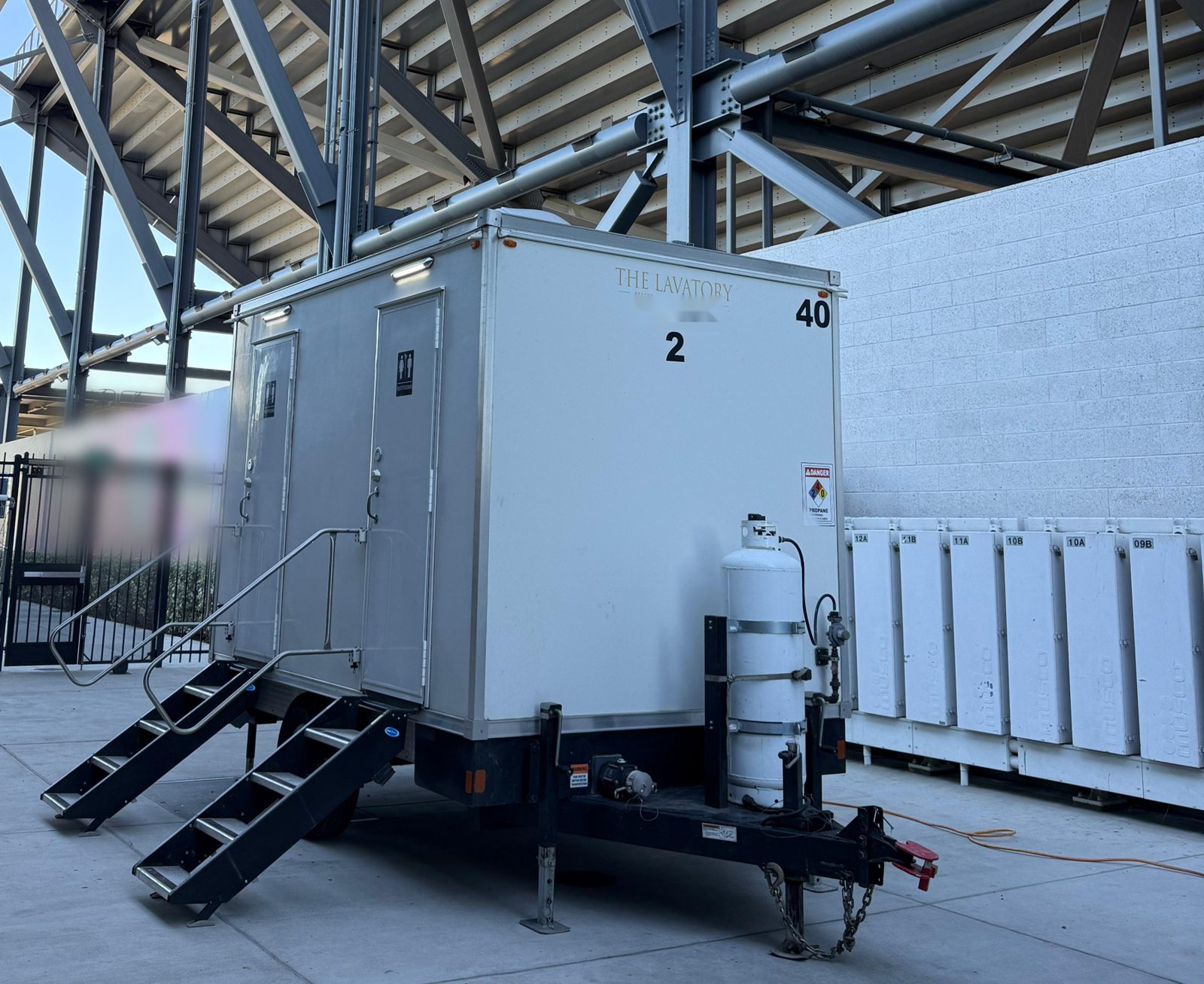 2-station shower and restroom combo trailer with two stairs, parked on concrete under the steel frame of a stadium grandstand