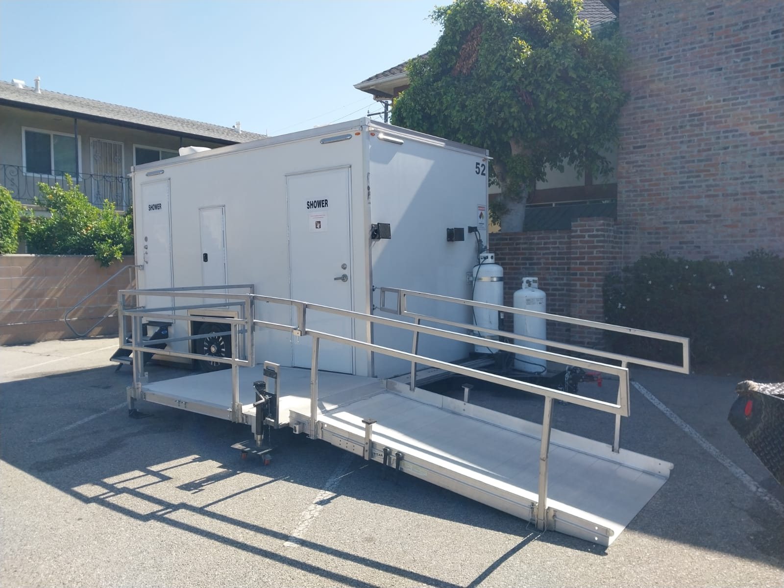 ADA plus 2 shower trailer with an aluminum ramp and railed landing, parked on a paved lot beside a brick building and a two-story residence hall style block