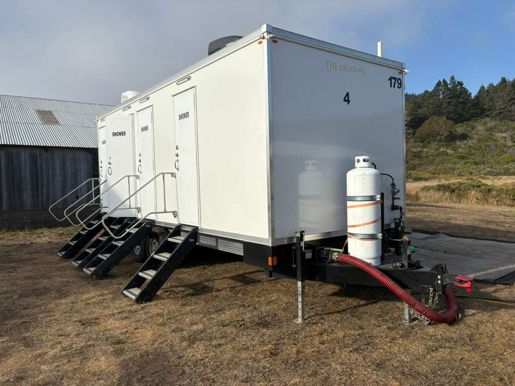 Multi-station shower trailer with SHOWER doors and railed stairs on a dry grass yard beside an old barn, with a red sewer hose run from the tongue