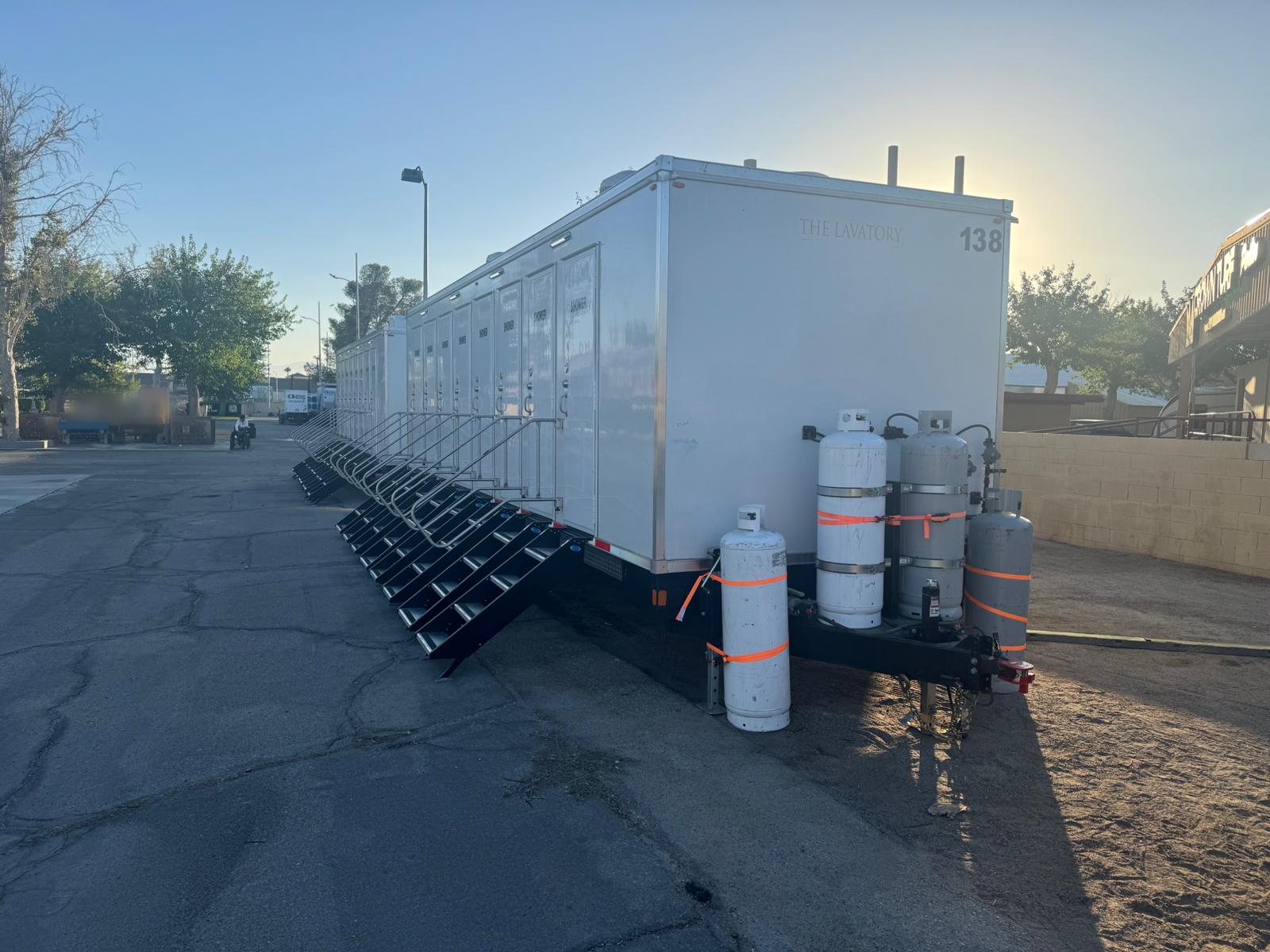 Two 8-station private shower trailers with SHOWER doors and black stair units lined up on a paved event lot at sunrise, propane tanks strapped at the tongue