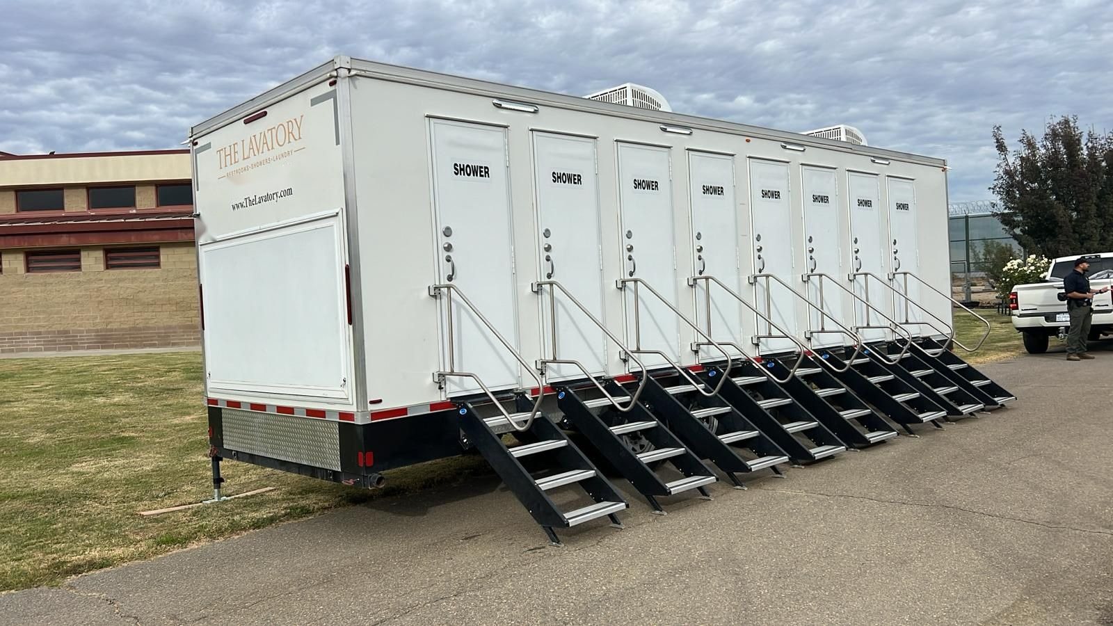8-station private shower trailer with eight stair units on a paved lot beside a block building, a staff member checking a clipboard next to a pickup