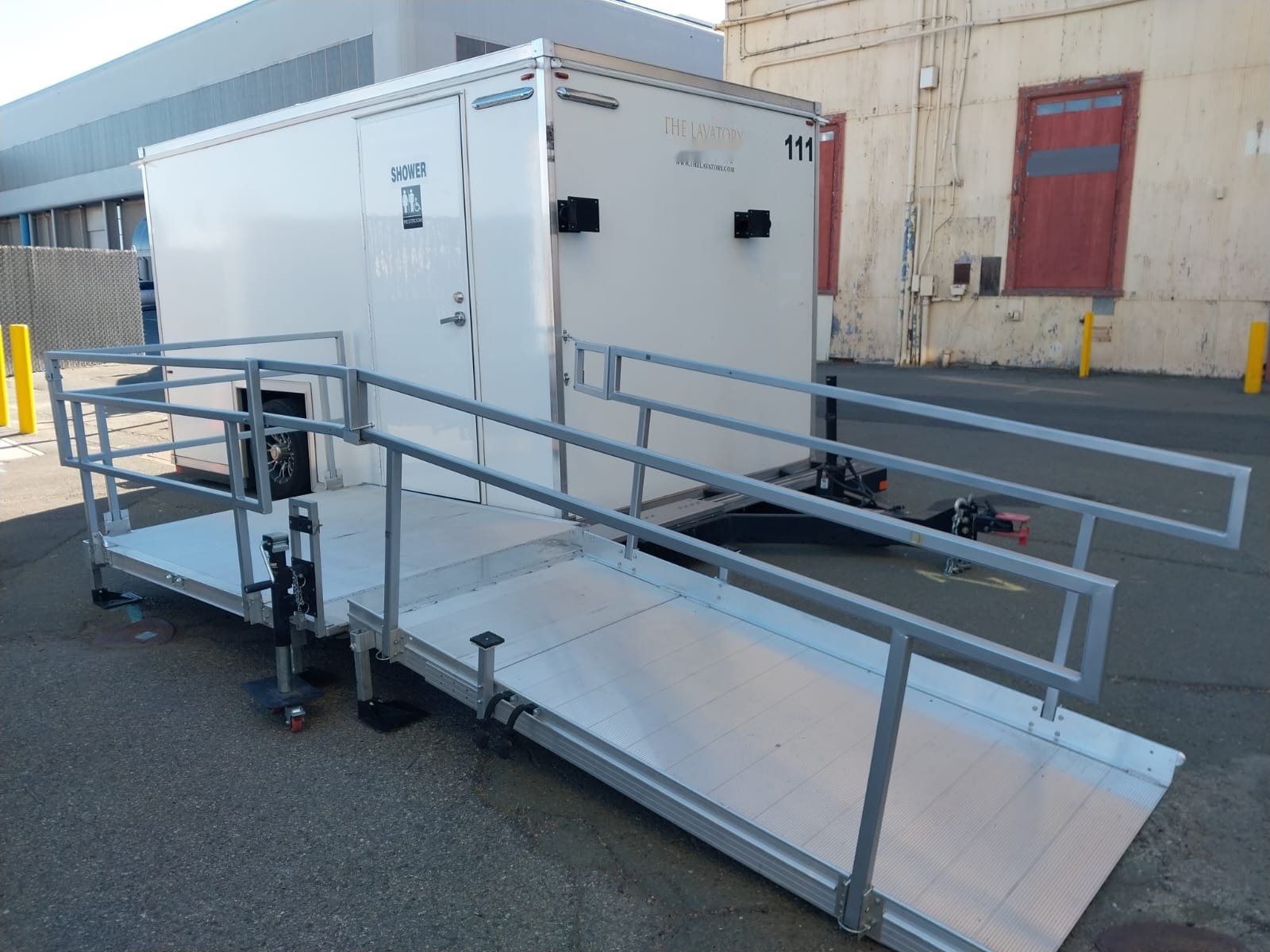 ADA + 2 shower trailer with its aluminum ramp and rails set up on a paved industrial lot between warehouse buildings