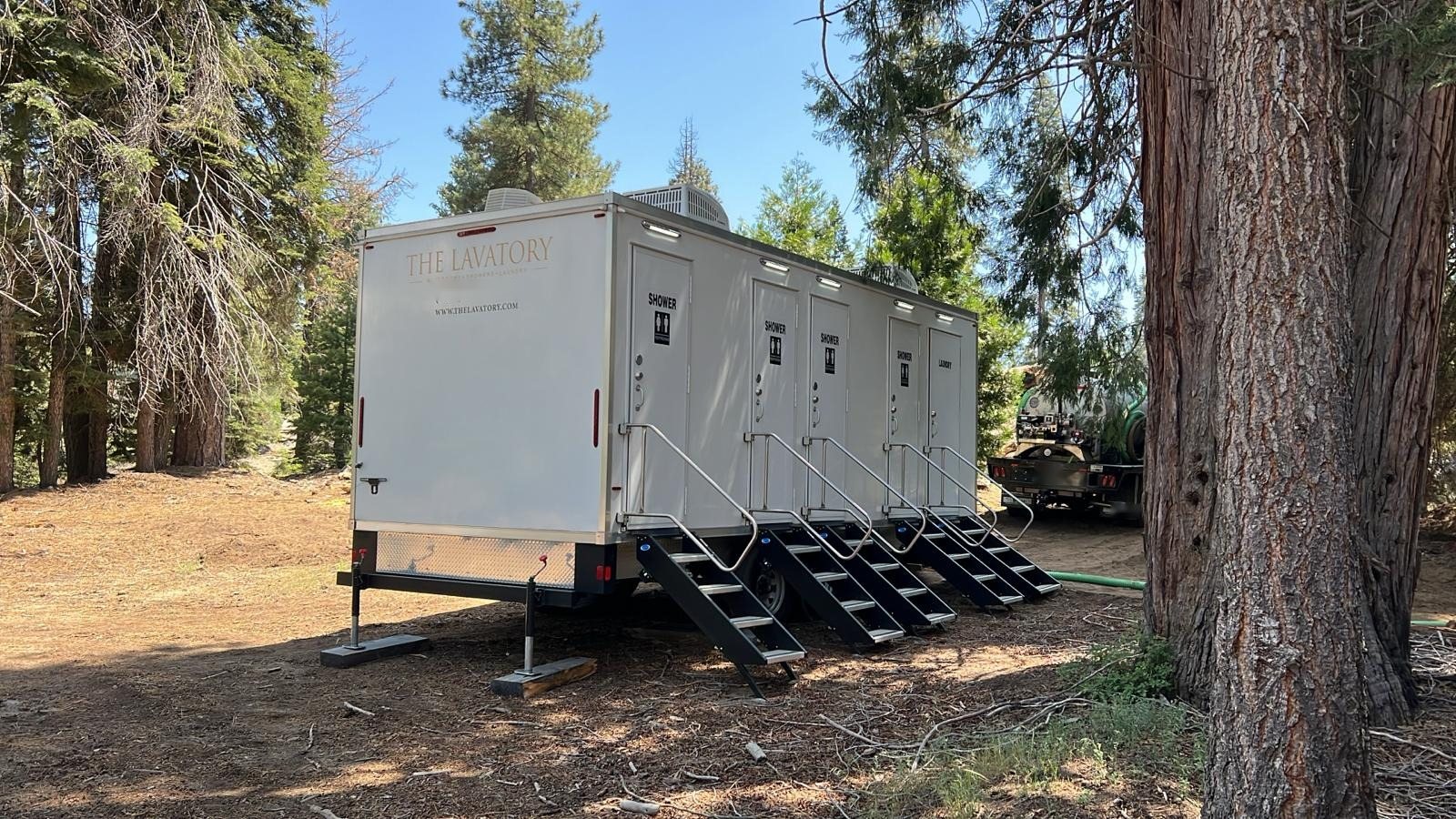 Shower and laundry combo trailer with four shower doors and a laundry door set on leveling pads in a shaded pine campsite, a service truck behind it