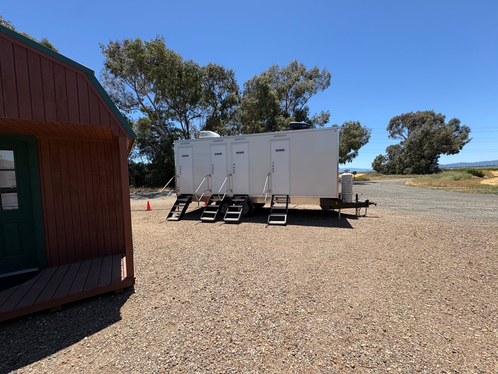 Private shower trailer with four doors and stairs on a gravel ranch yard beside a red barn-style shed and tall eucalyptus trees