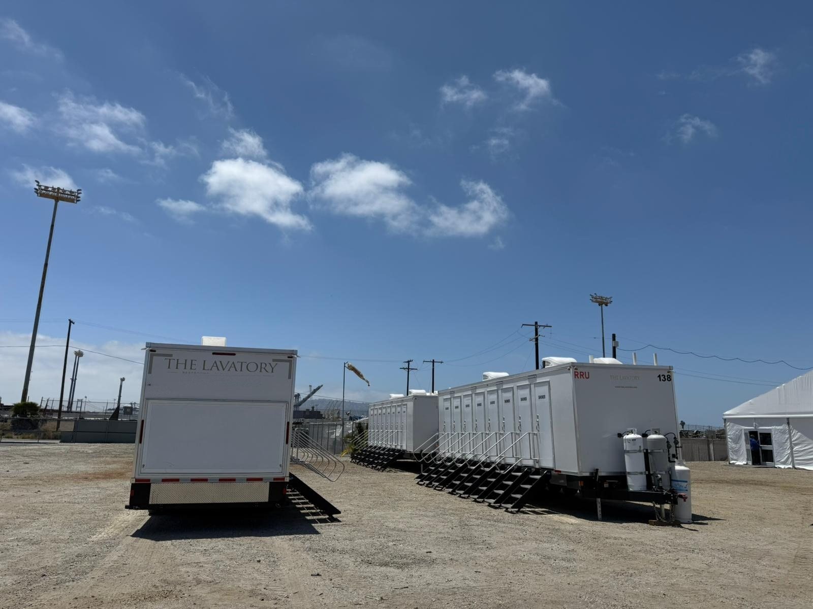 Three shower trailers staged on a gravel yard beside a white tent, with power poles and light towers behind them