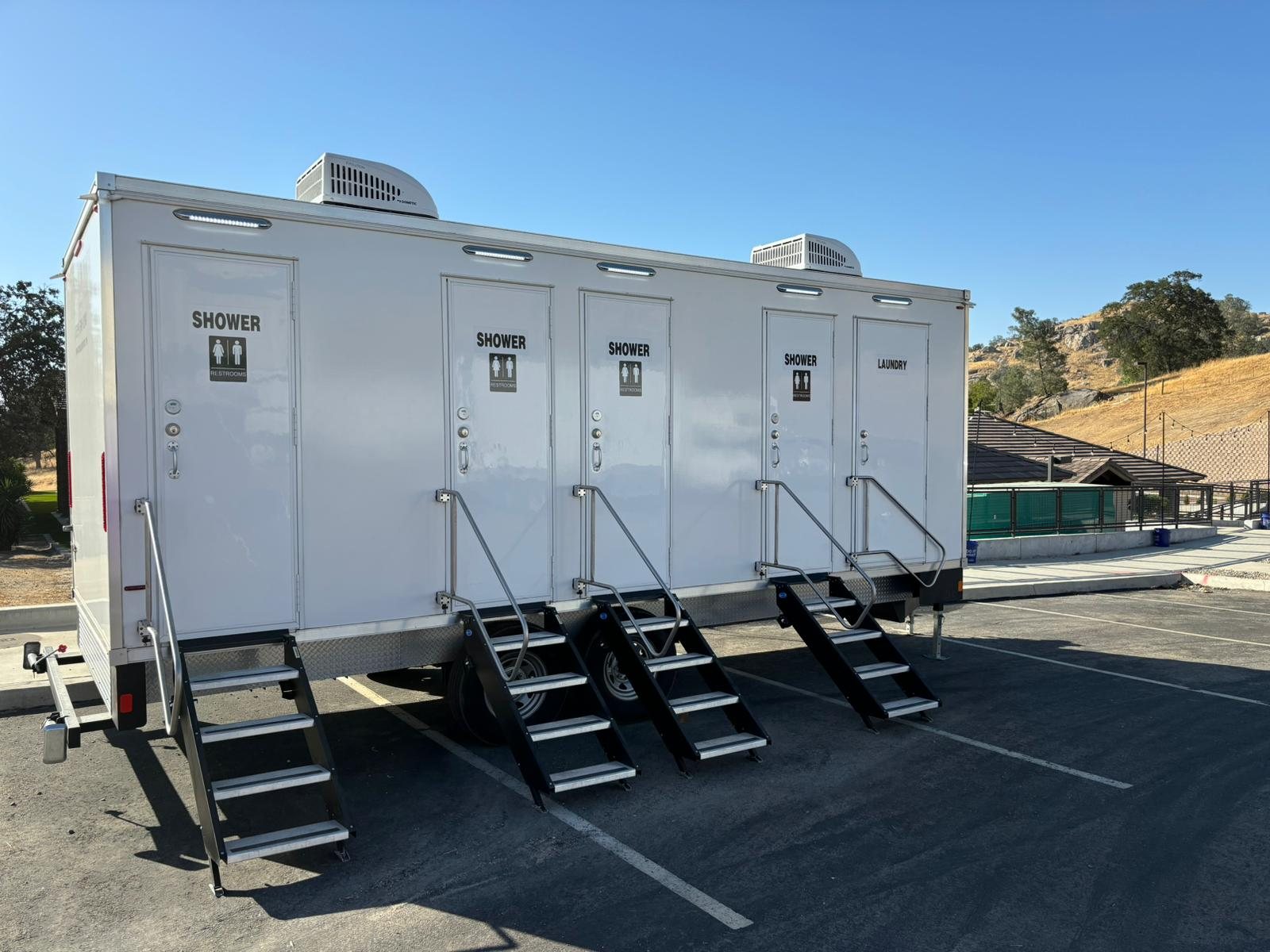 4-station shower, restroom and laundry combo trailer on a paved lot with stair units at each door and dry hills behind it