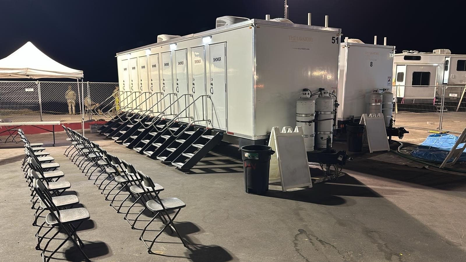 8-station shower trailer lit up at night at a fenced base camp, folding chairs in rows and two crew members standing by a canopy