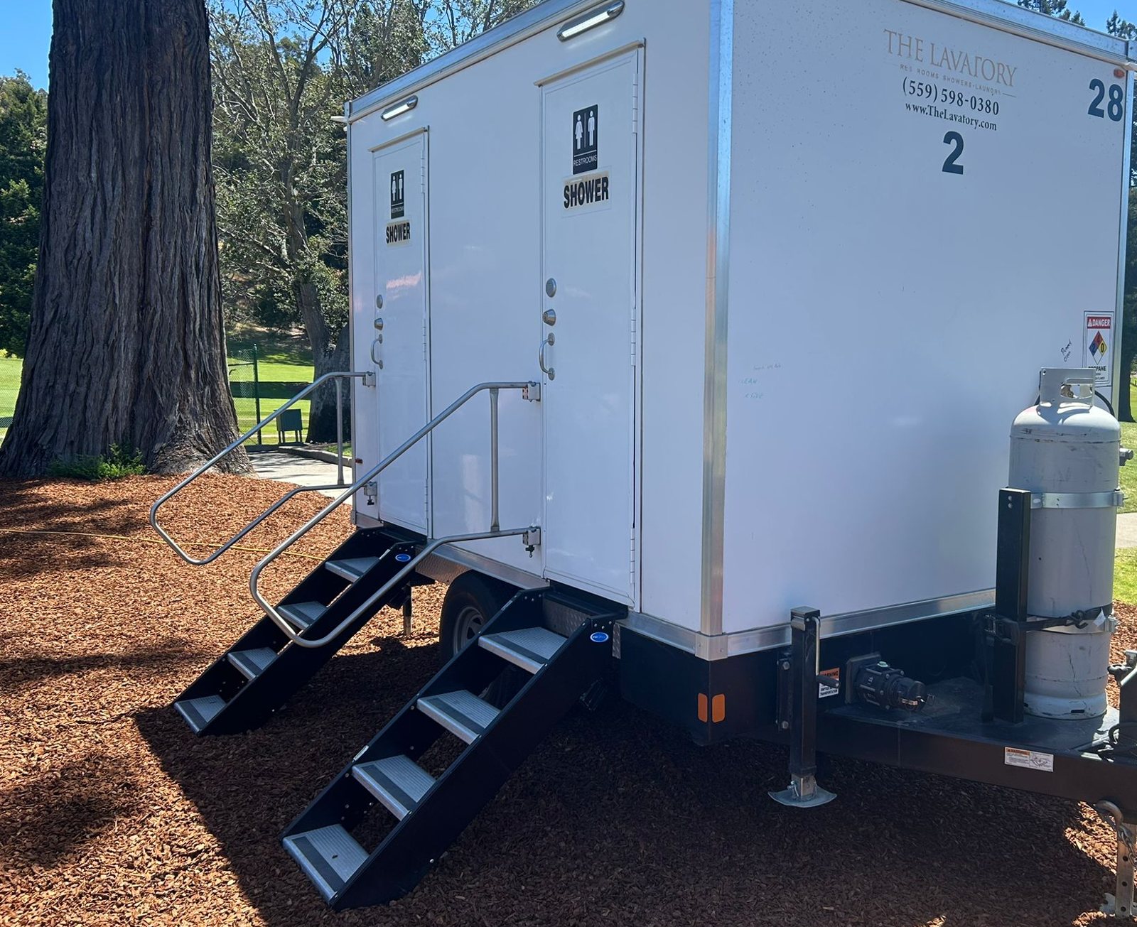 2-station shower and restroom combo trailer set on wood mulch under a large tree at the edge of a green lawn