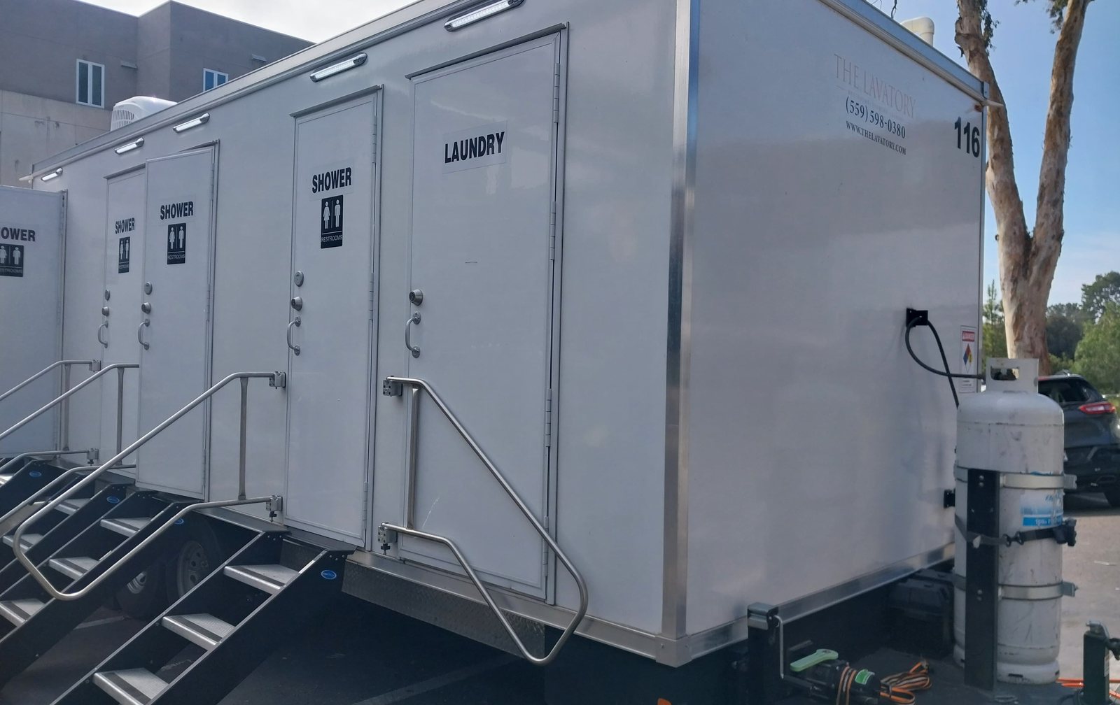 Shower, restroom and laundry combo trailer with shower doors and a laundry door, parked on a lot beside an office building and a eucalyptus tree