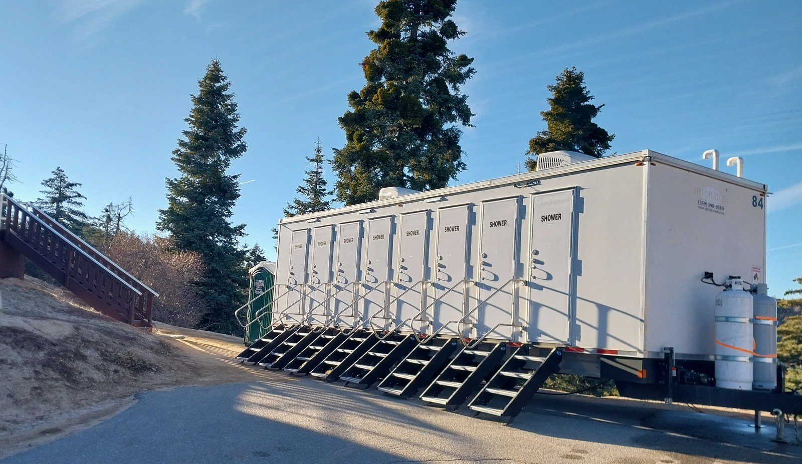 8-station private shower trailer with a stair at each of its eight doors, parked on a paved drive below tall pines and a wooden stairway