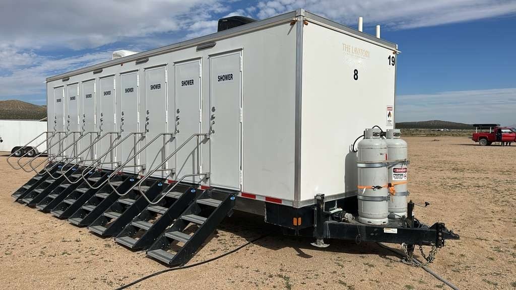 8-station shower trailer with eight stair units set on a dirt staging lot, a work truck parked in the distance