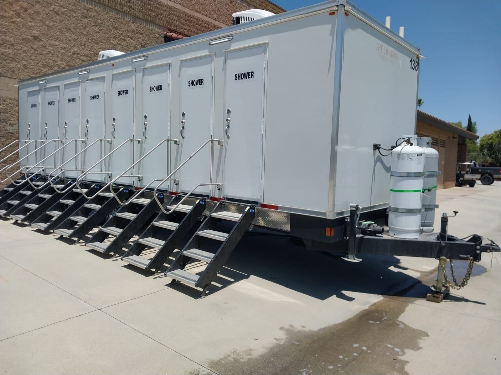 8-station private shower trailer with eight doors and eight stair units parked on a concrete lot beside a block building, propane tanks on the tongue