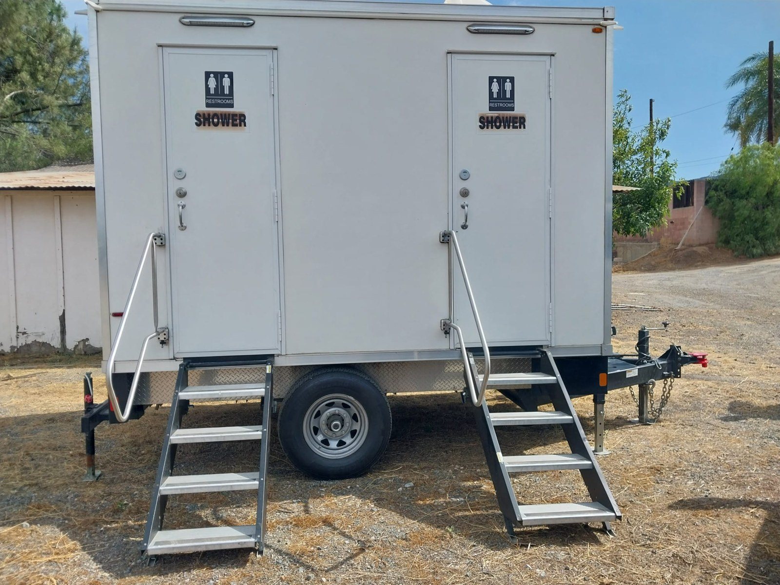 2-station shower and restroom combo trailer on a dry dirt yard beside a farm shed, two doors with their own stairs