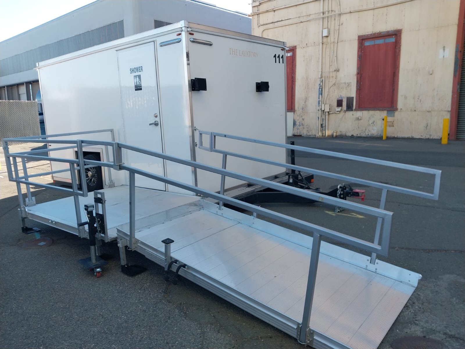 ADA shower trailer with an aluminum ramp, landing and handrails set up on a paved industrial yard beside an old warehouse building