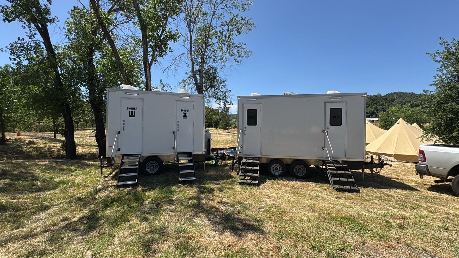Two-room shower trailer and a restroom trailer parked side by side on a grassy field under shade trees, with canvas camp tents beside them