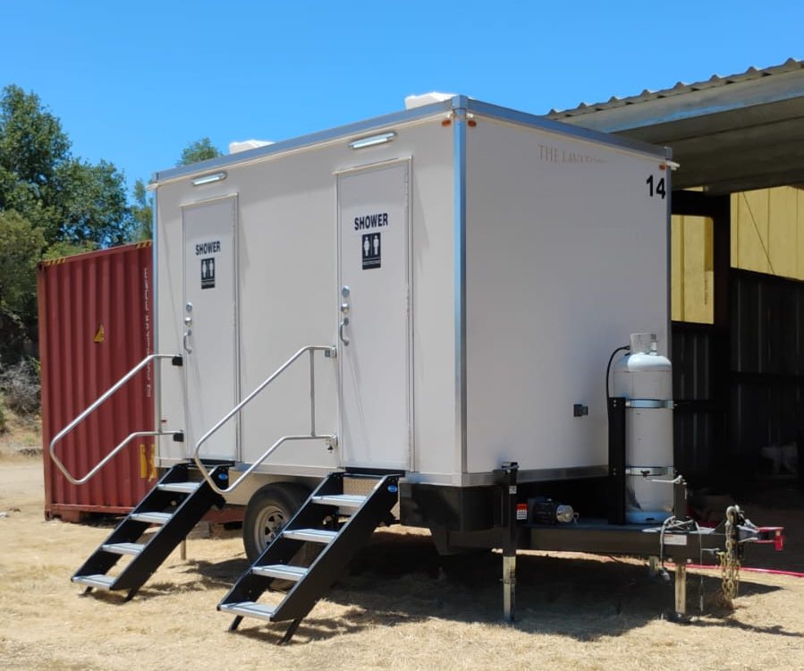 2-station shower trailer on dry grass beside a red shipping container and an open equipment shed on a rural property