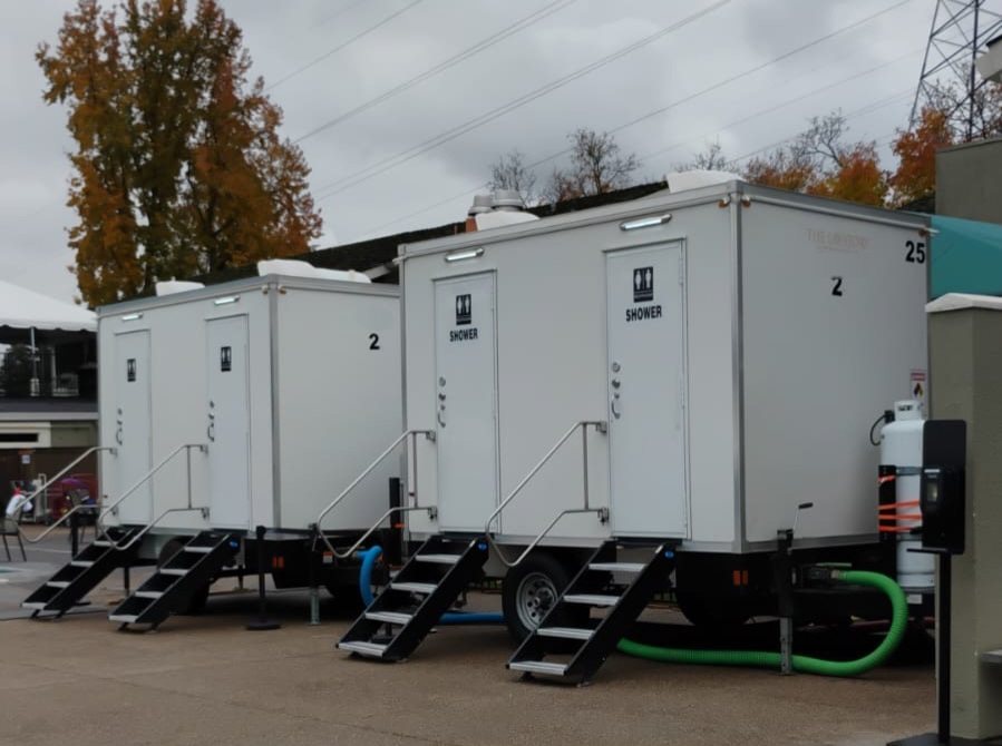 Two 2-station shower trailers with stairs and hoses connected, parked on a concrete lot under high-voltage power lines on a cloudy day