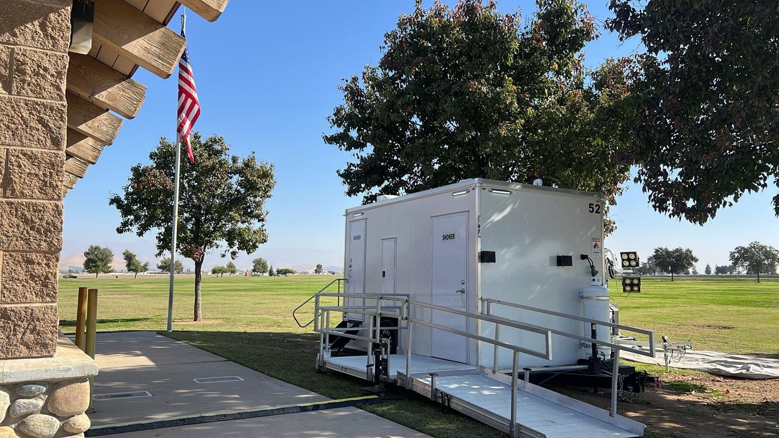 ADA plus two shower trailer with a ramp and rails set beside a stone public building and a flagpole on a park lawn