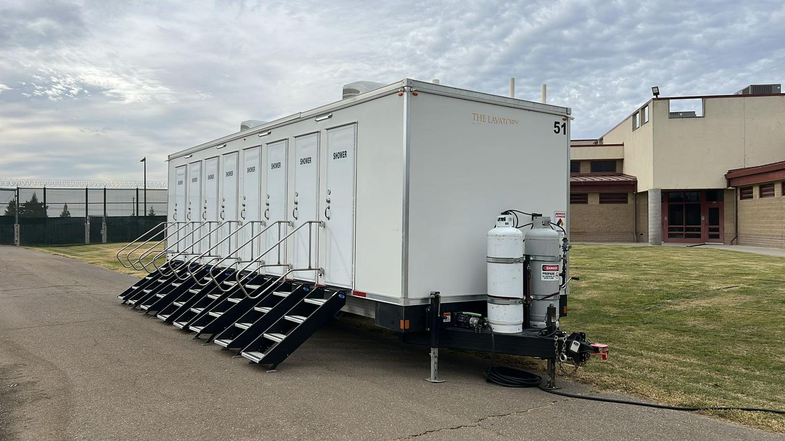 8-station private shower trailer with eight doors and eight stair units parked on a paved drive beside a lawn and a facility building